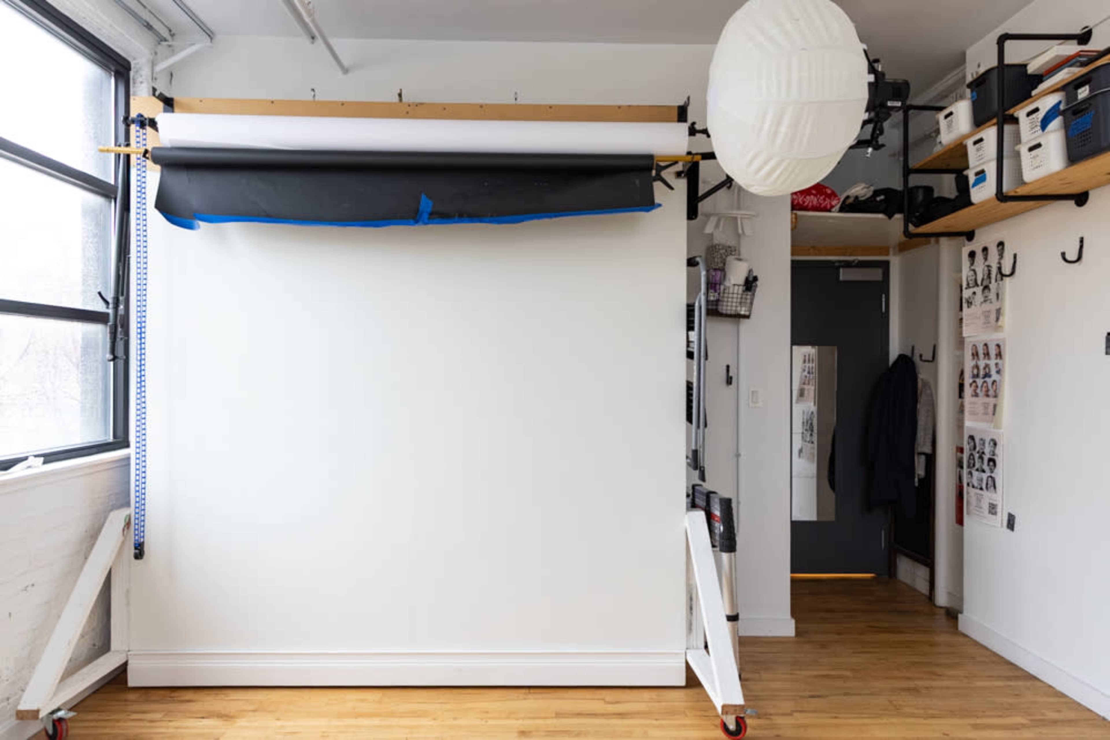 A photography studio features a white backdrop mounted on a support system, with storage boxes and a lamp in the surrounding area.