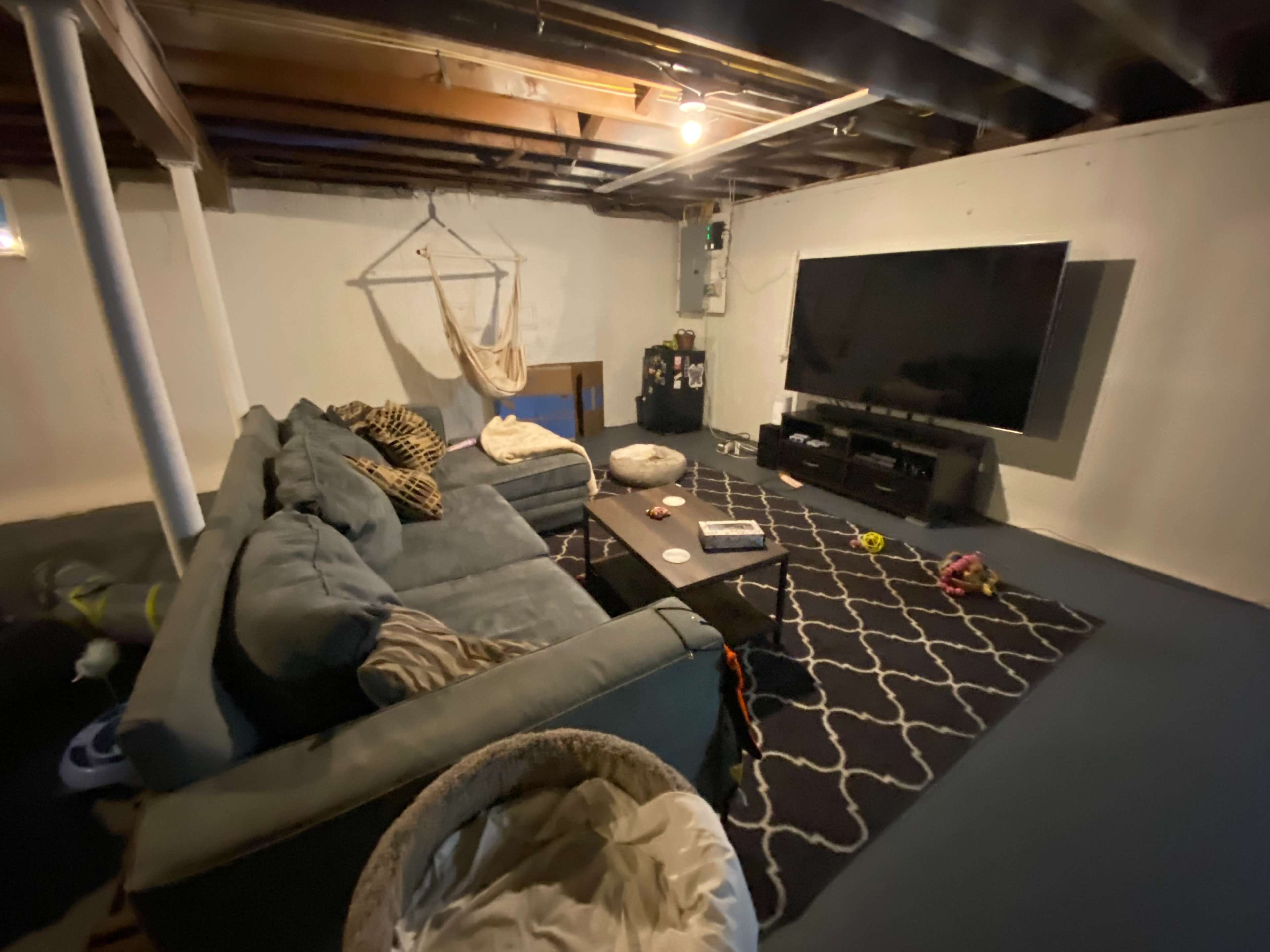 A basement living area featuring a large sectional sofa, a coffee table, and a wall-mounted television against a white wall.