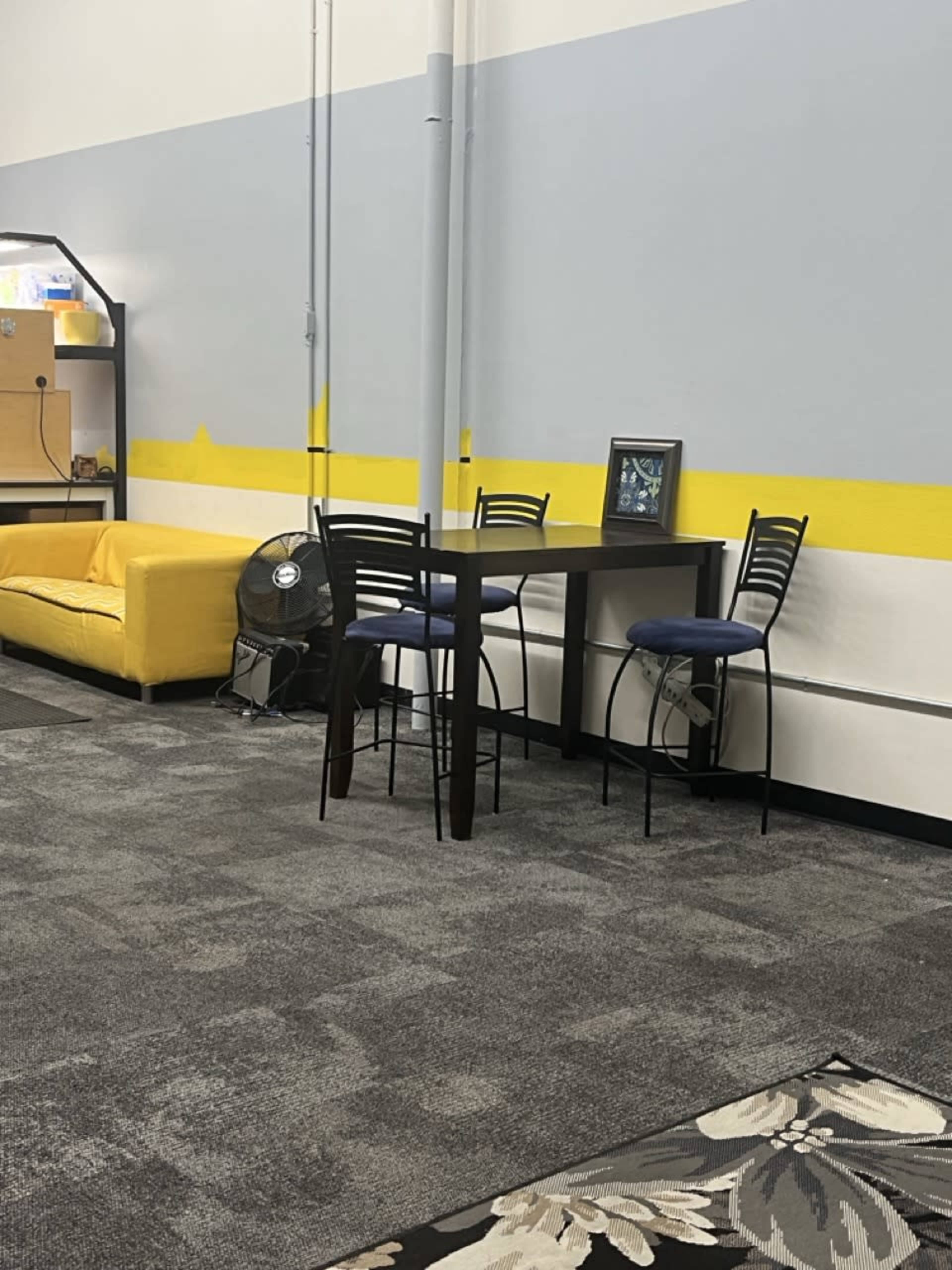 The image shows a sparse indoor space featuring a yellow couch, a small table with four black chairs, and a fan against a background of gray walls with a yellow stripe.