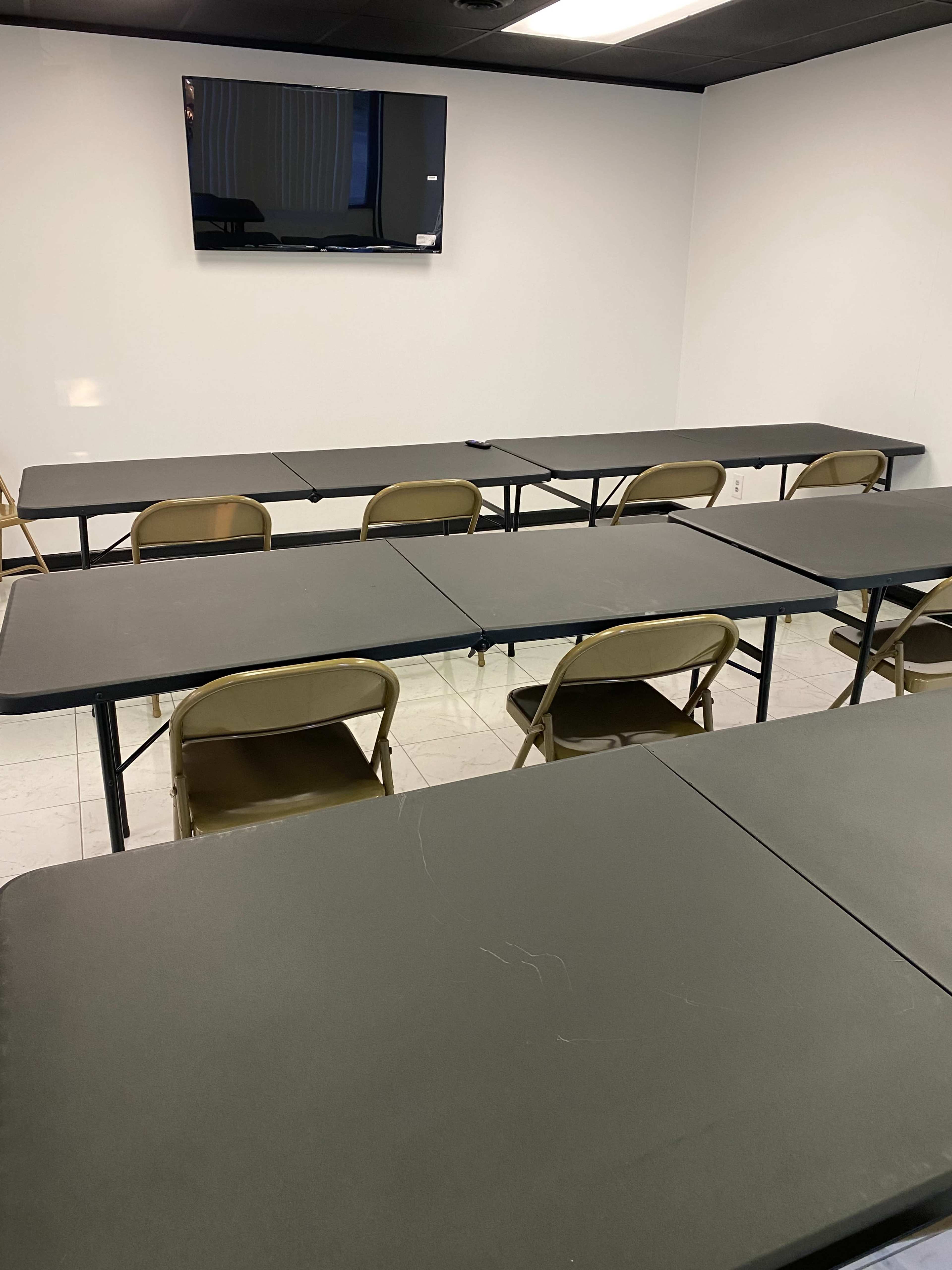 A classroom or meeting room with several black tables arranged in rows and foldable chairs, along with a flat-screen TV mounted on the wall.