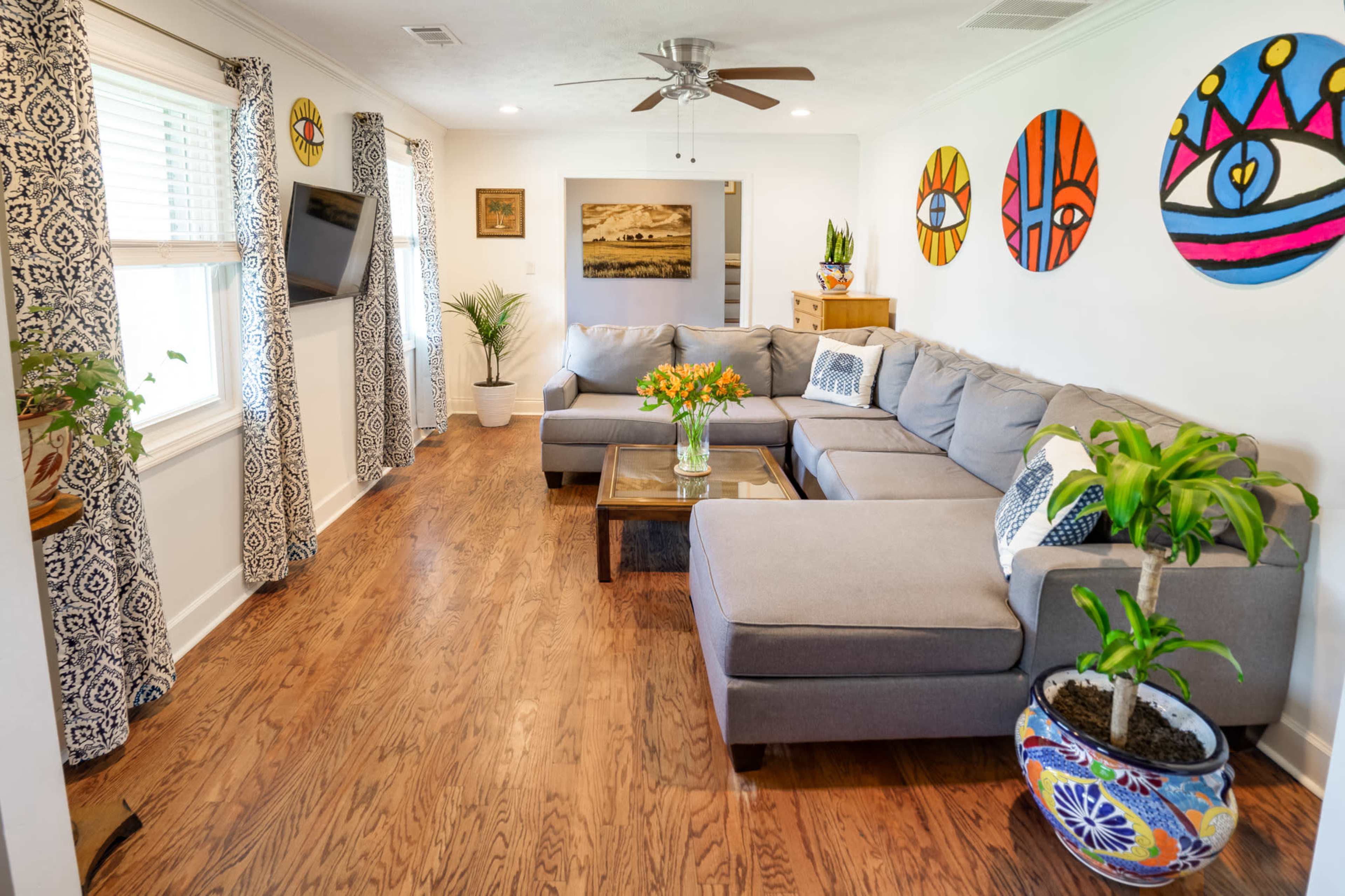 A bright living room featuring a gray sectional sofa, a wooden coffee table with a bouquet of flowers, and colorful wall art.