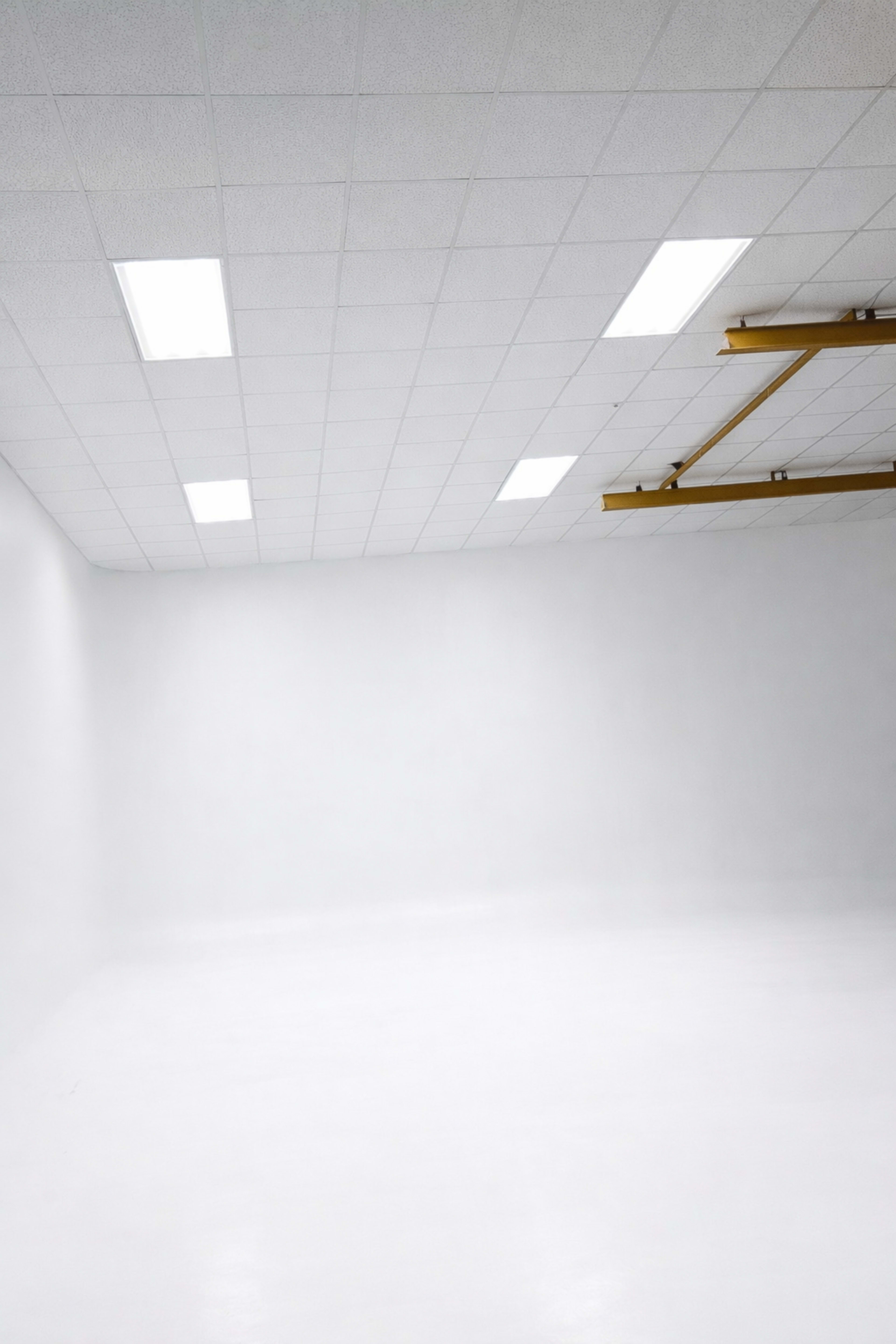 The image shows a spacious, empty room with white walls and a flat ceiling illuminated by fluorescent lights.