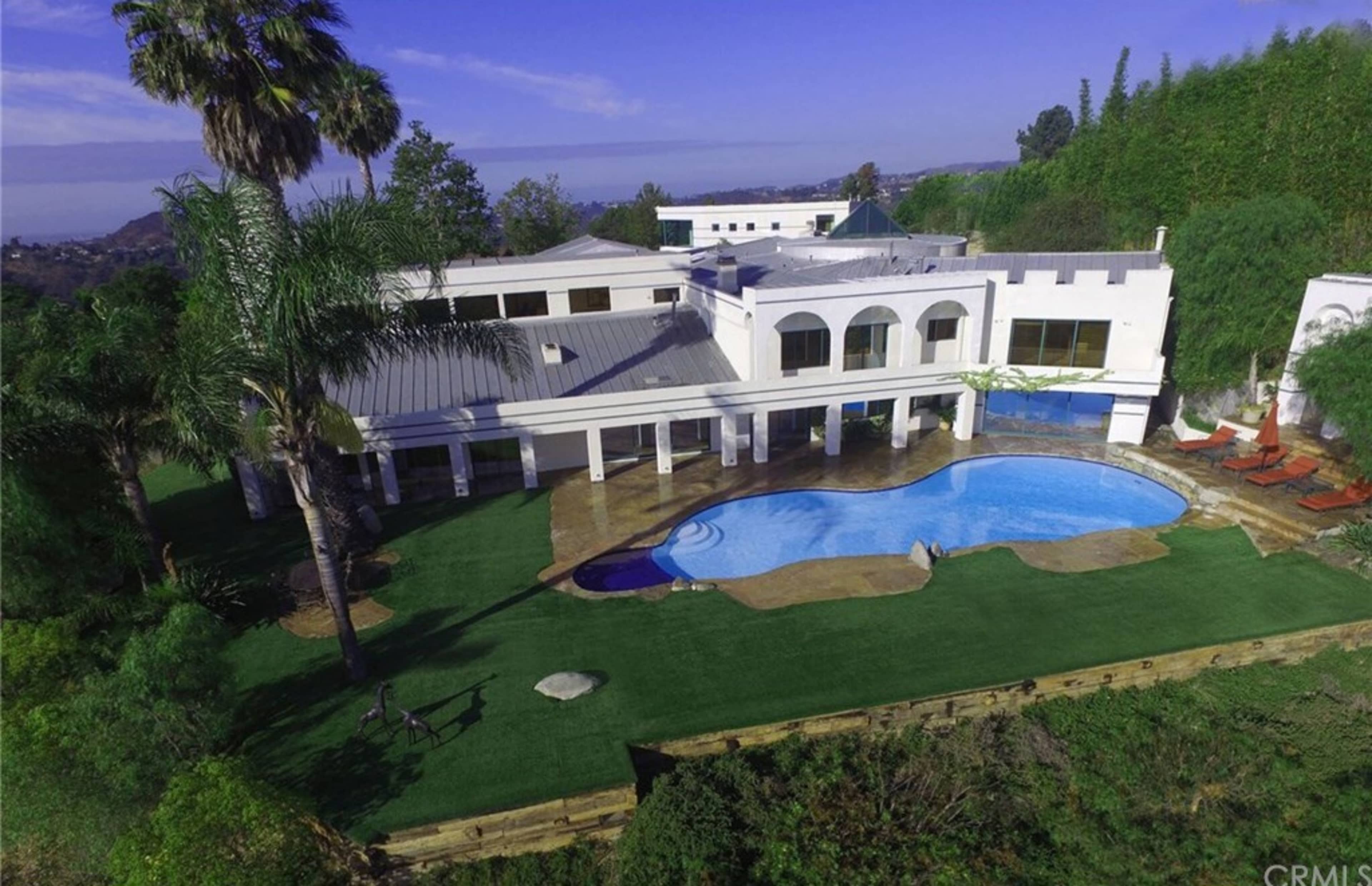 A large house with a swimming pool, surrounded by landscaped greenery and palm trees.