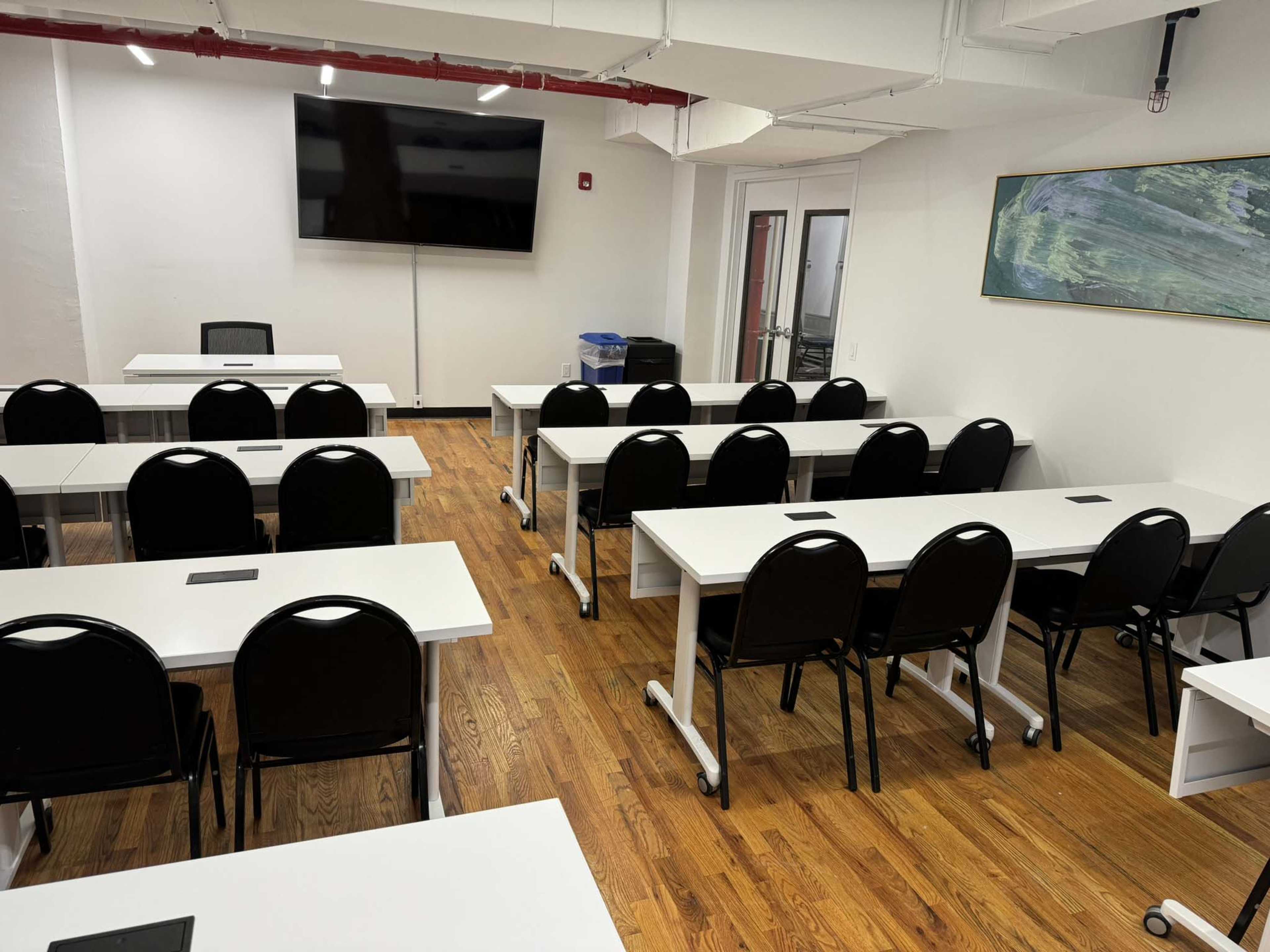 A classroom with white tables arranged in rows, black chairs, a wall-mounted screen, and a large window.