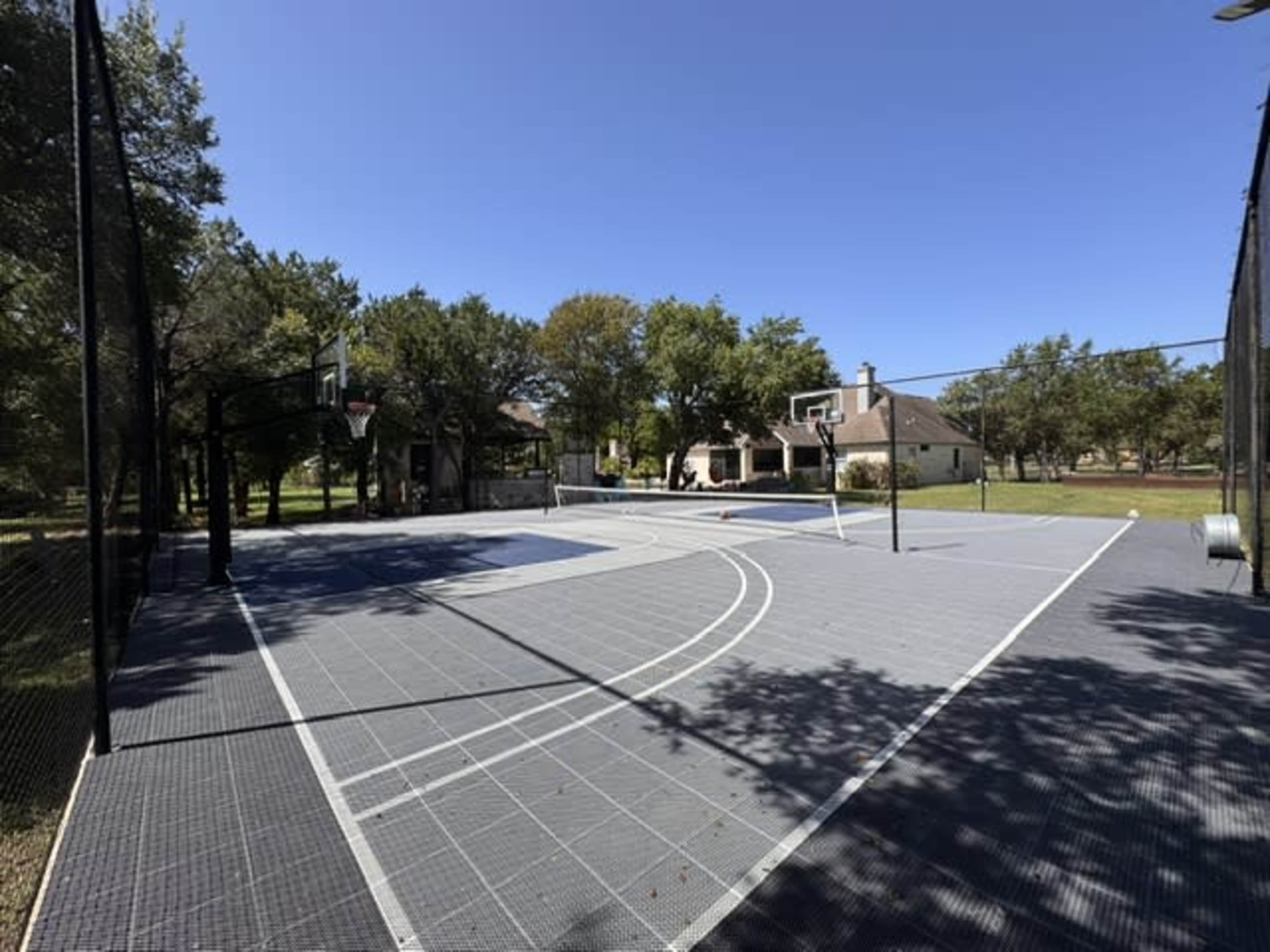 The image shows a fenced outdoor basketball court with multiple hoops, surrounded by trees and a residential building in the background.