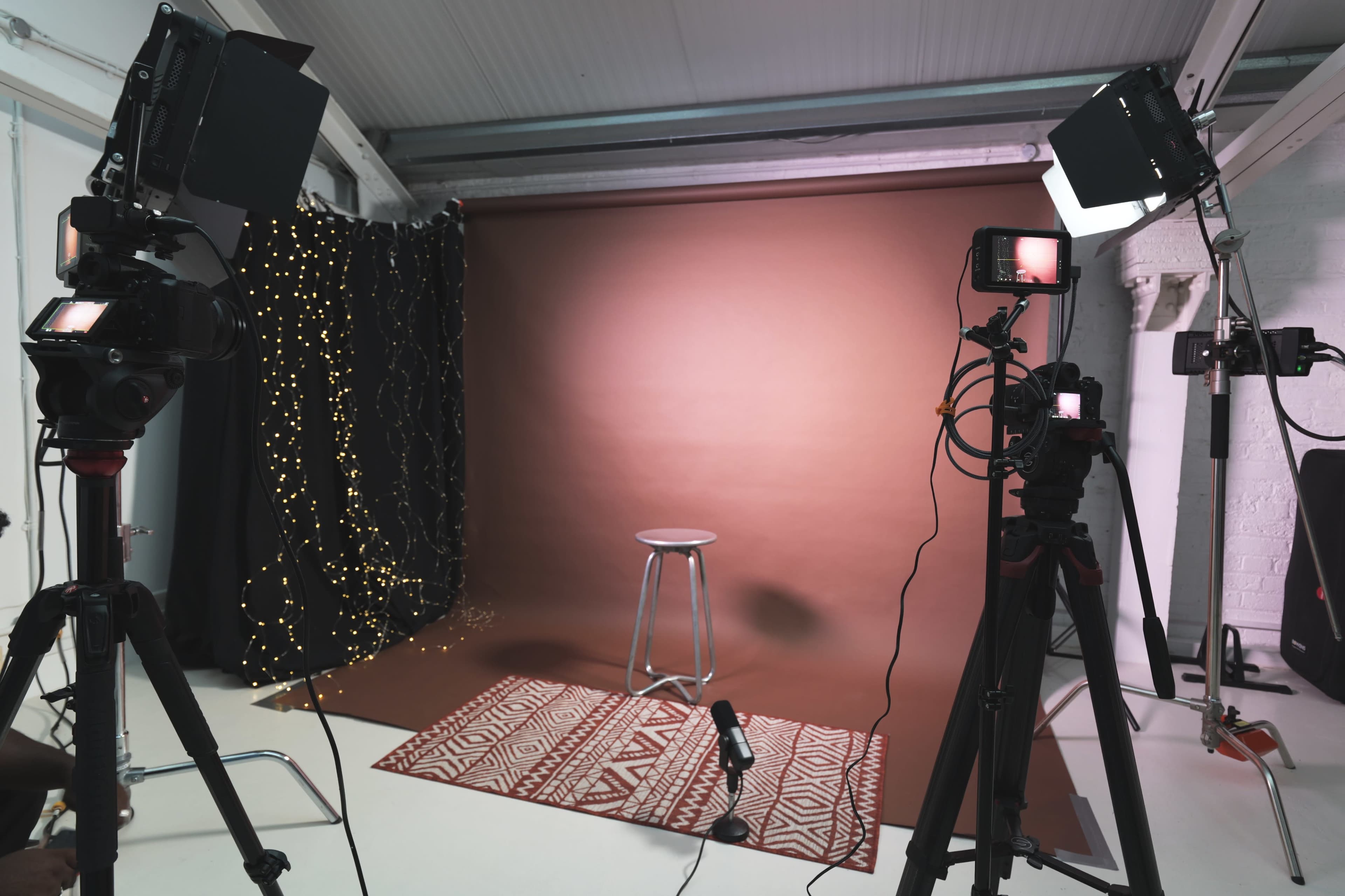 The image shows a photo studio setup with cameras, lighting equipment, and a stool on a patterned rug against a brown backdrop.