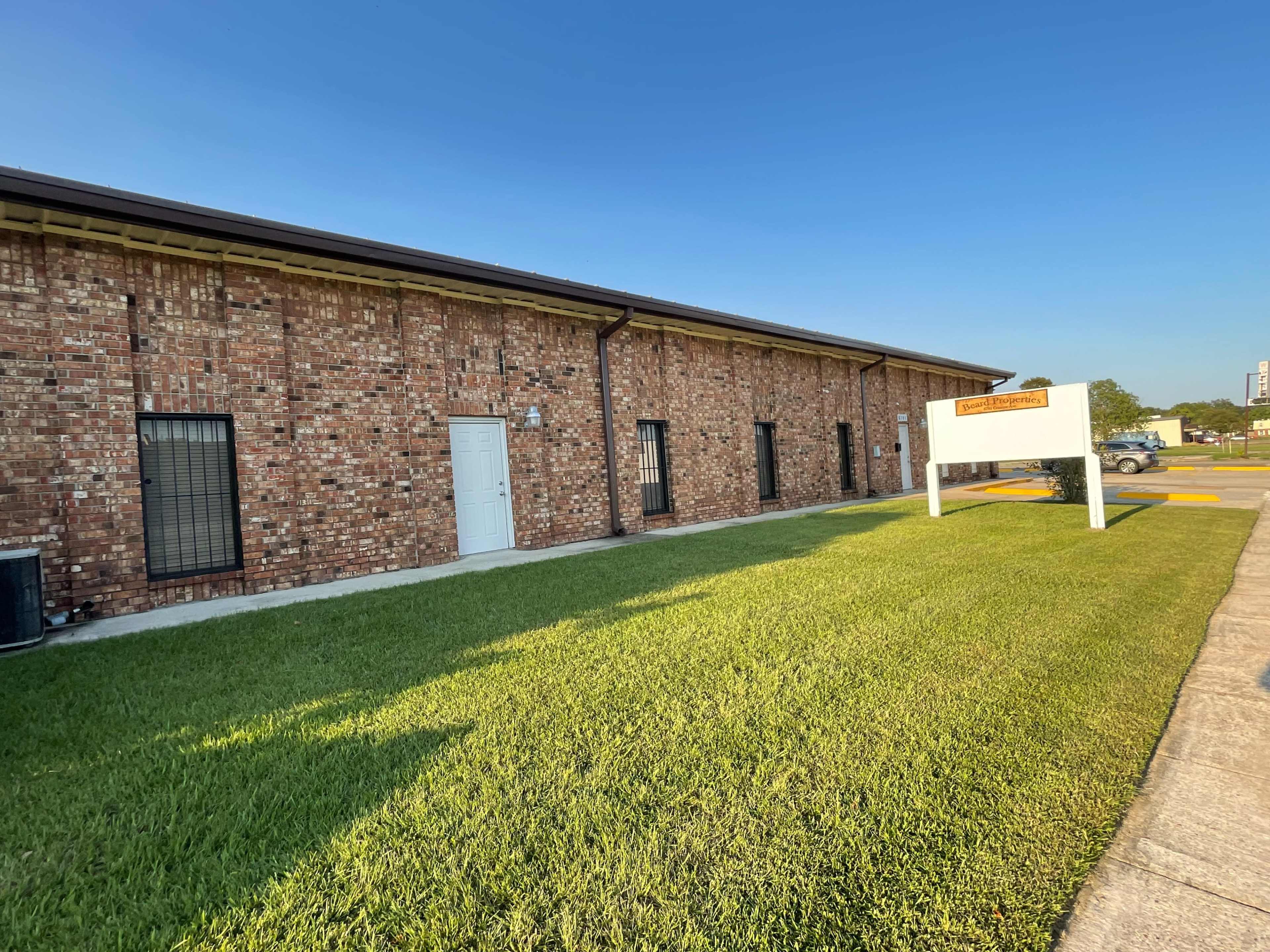 A brick building with a white door and windows is surrounded by neatly manicured grass and a sign in front.