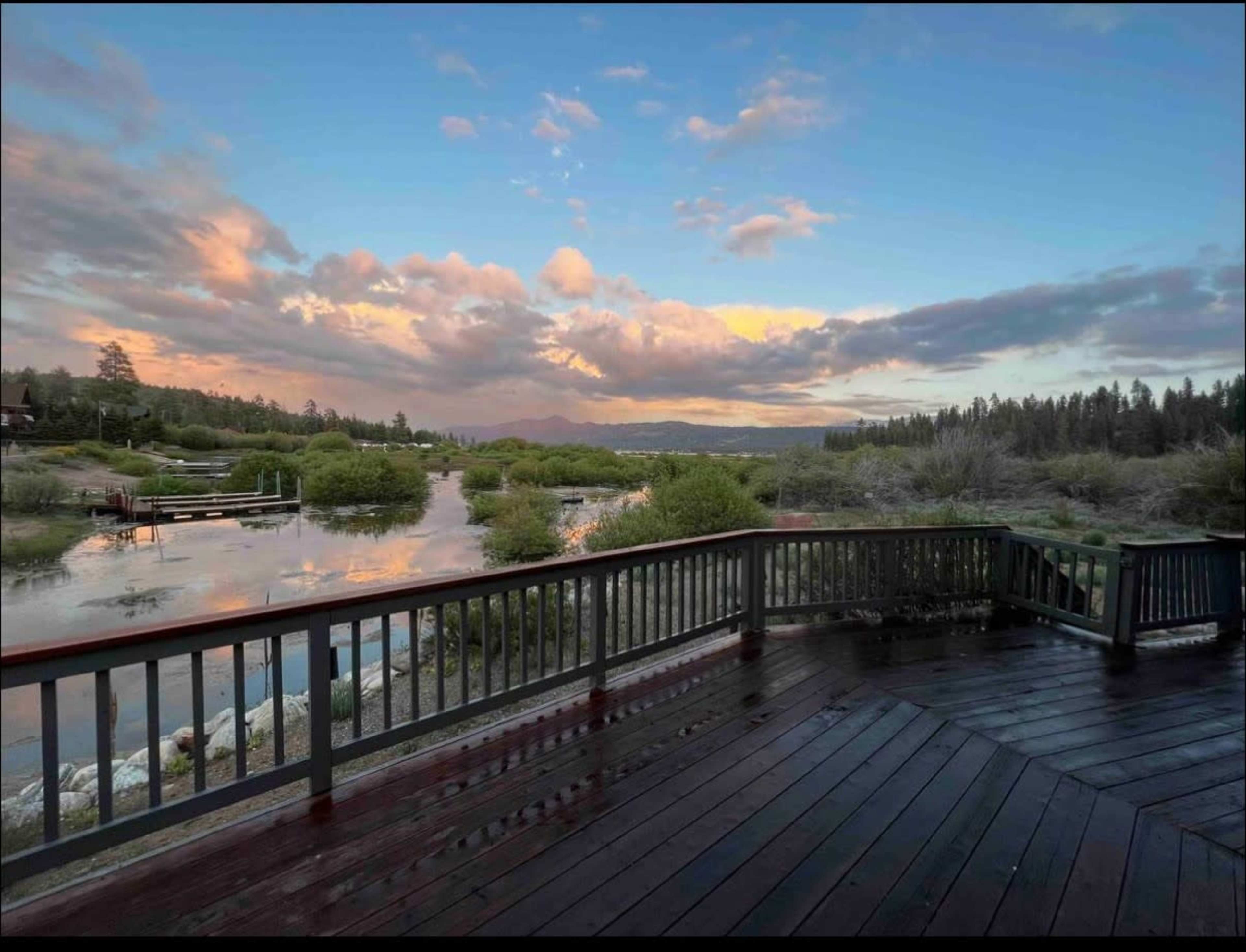 The image shows a scenic view from a wooden deck overlooking a calm body of water surrounded by greenery and clouds in the sky.