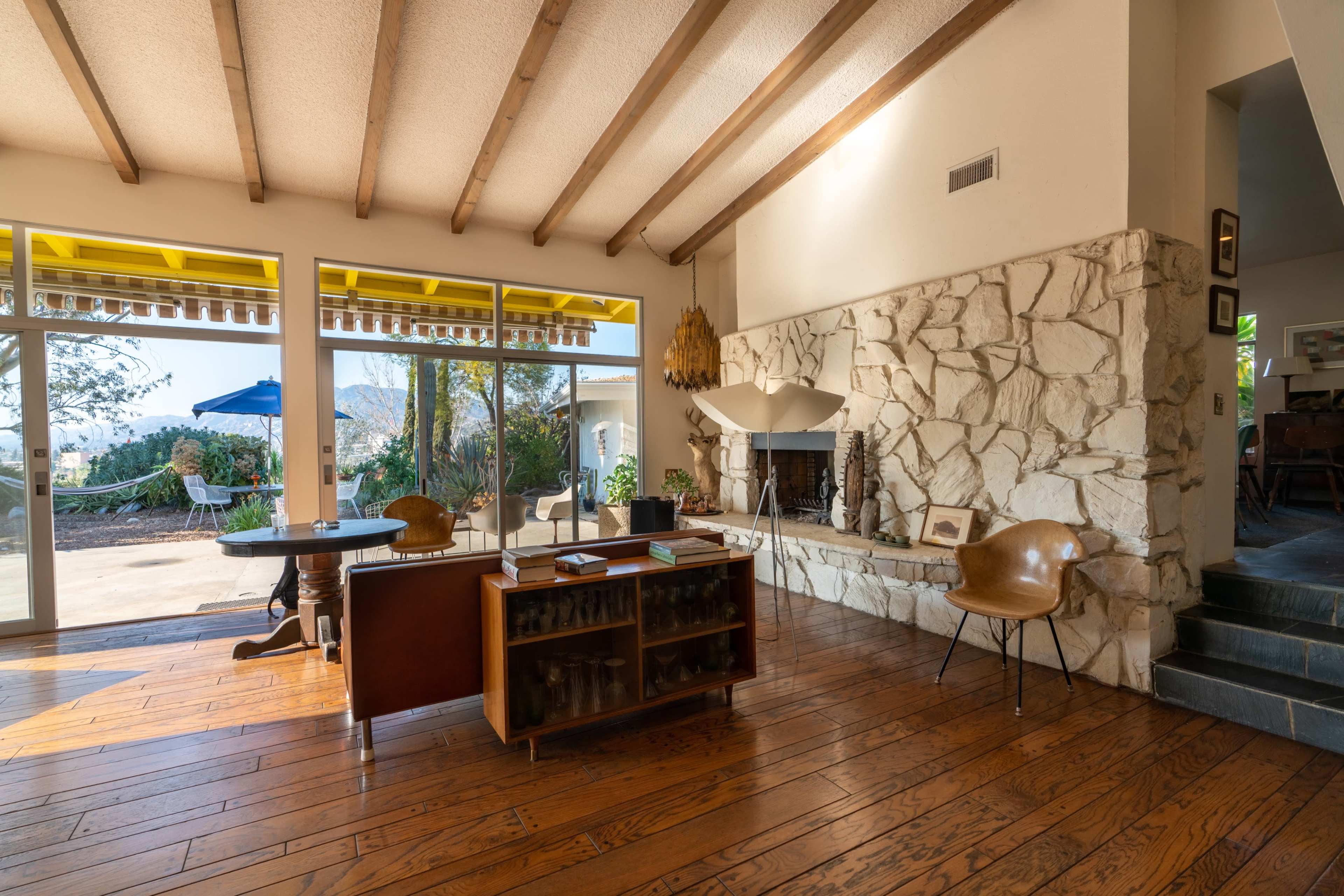A modern living room with wooden beams, a stone fireplace, and a view of an outdoor patio through large windows.