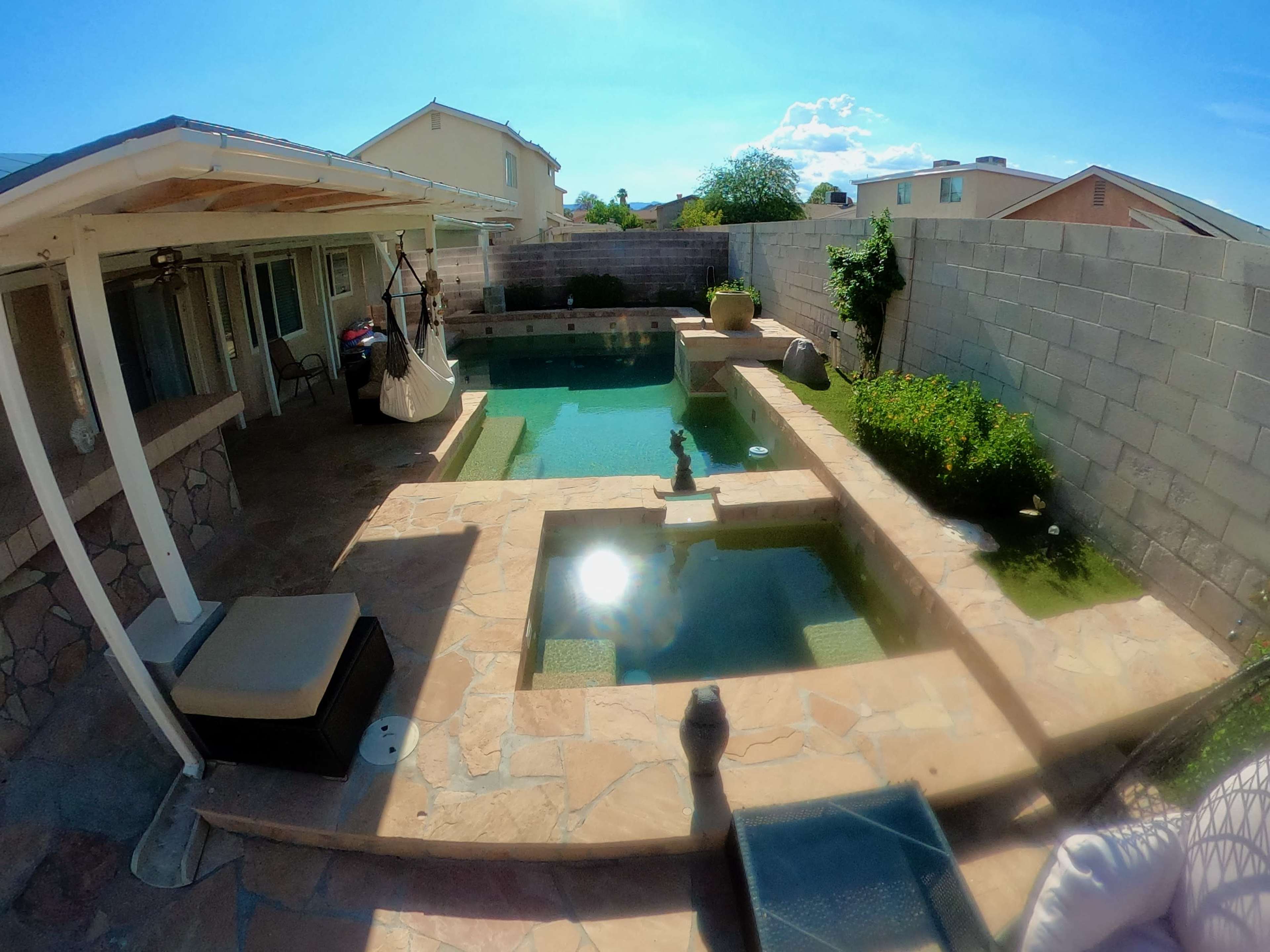 A backyard pool area with a stone patio, featuring a rectangular pool, a small garden, and a covered seating area.