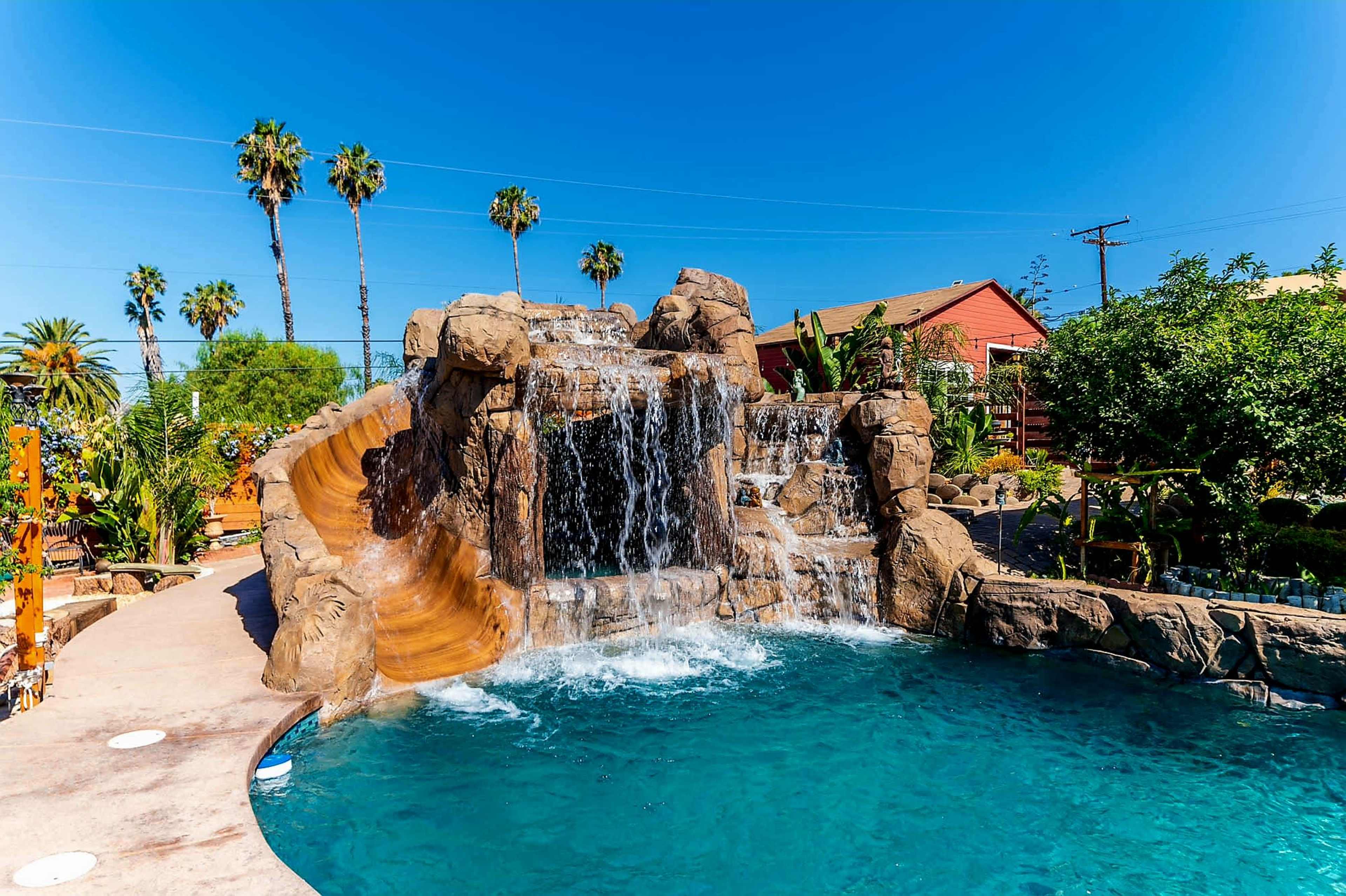 The image shows a swimming pool with a rock formation and waterfall, surrounded by palm trees and a house in the background.