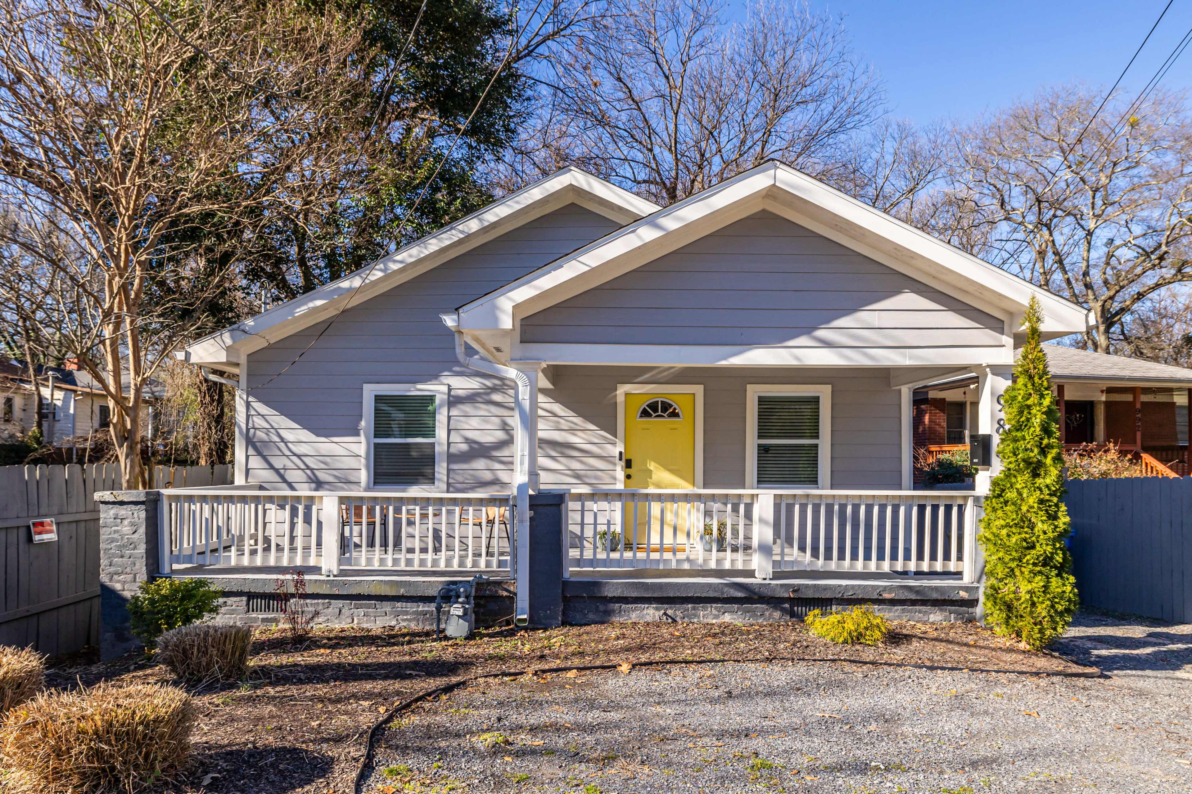 A single-story gray house with a yellow front door and a porch is surrounded by a gravel driveway and a low stone wall.