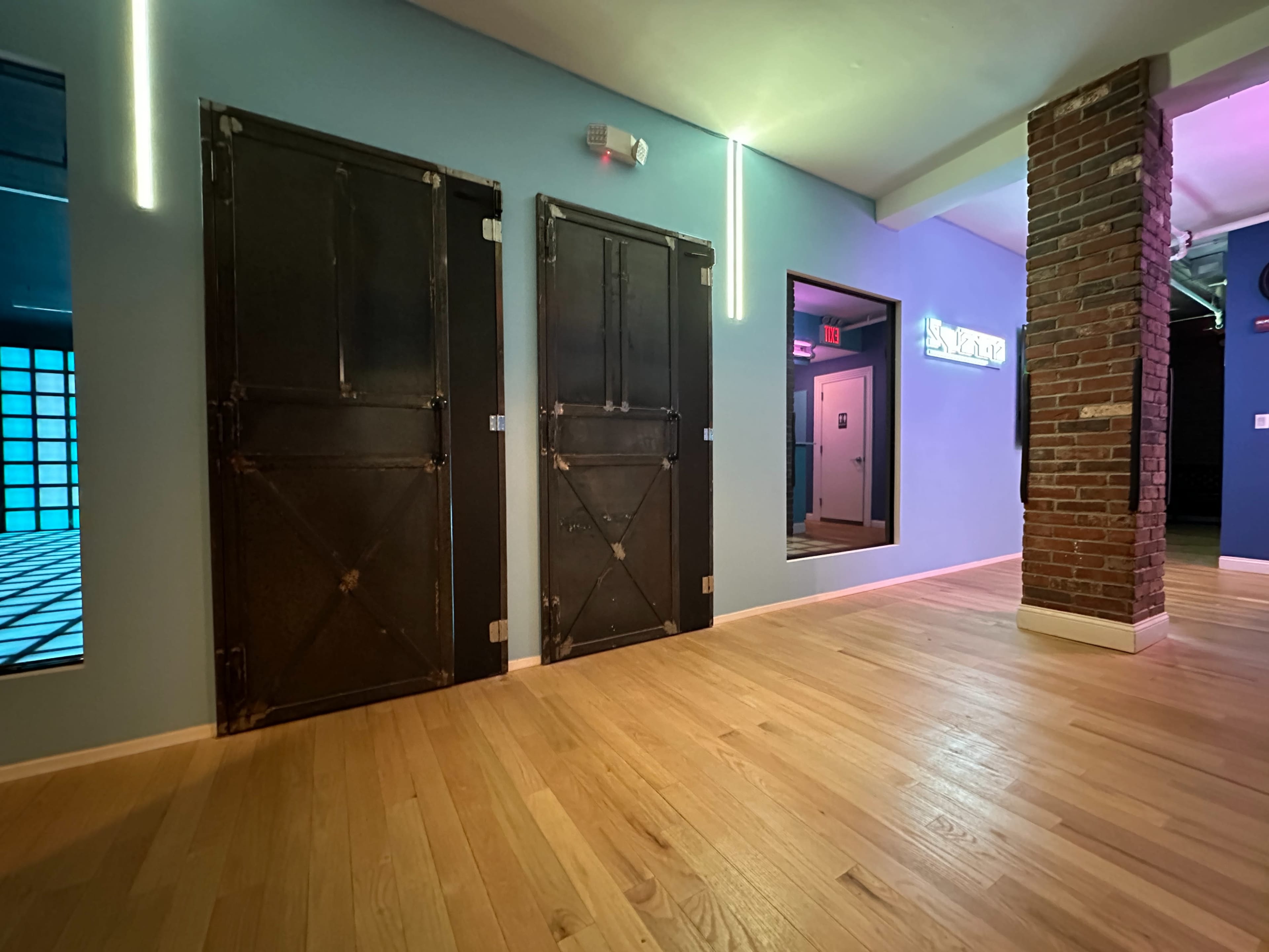 A hallway with two dark metal doors on the left, a brick column on the right, and a brightly lit room in the background.