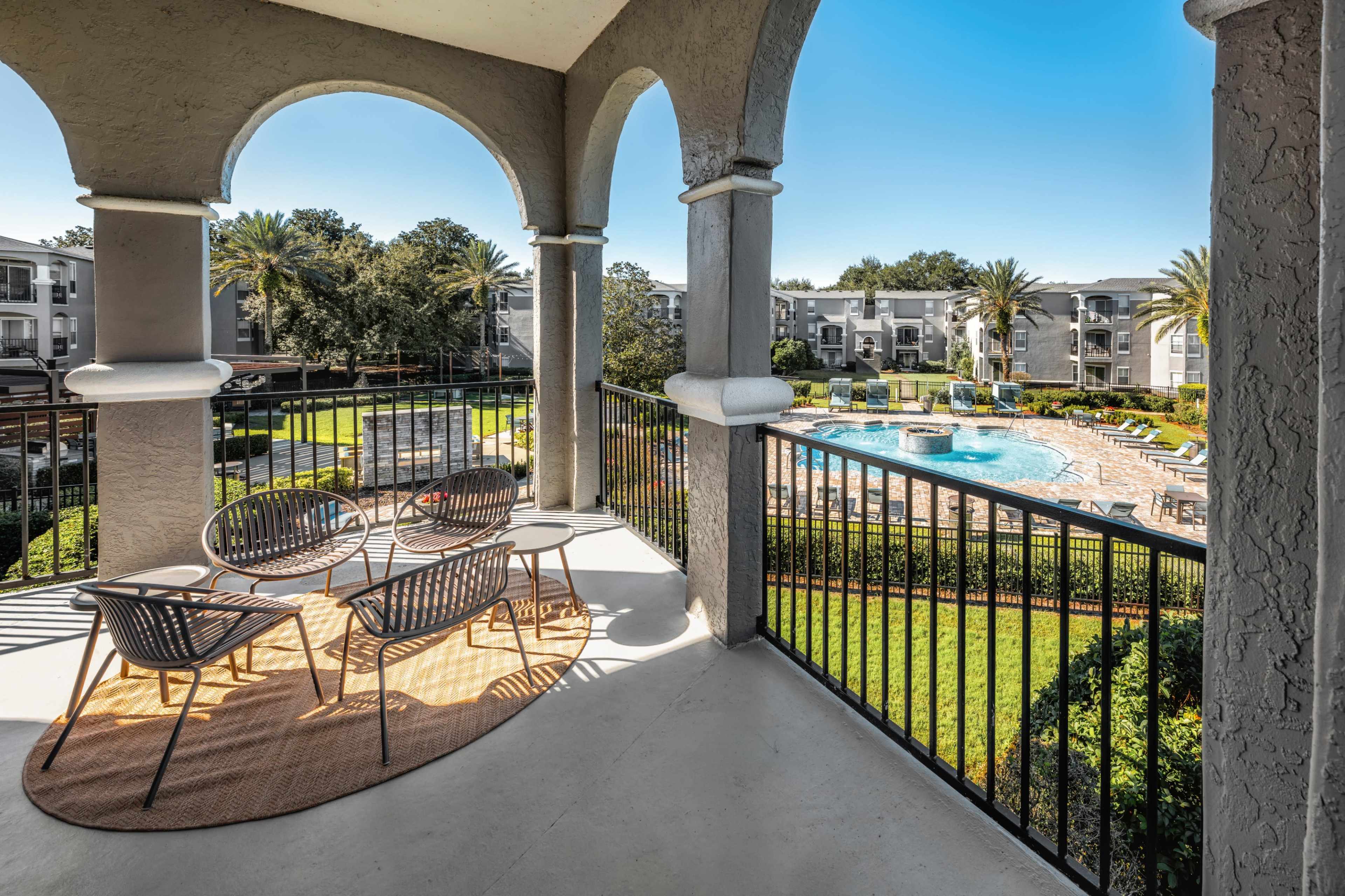 A covered balcony with modern chairs overlooks a resort-style pool surrounded by palm trees and landscaped gardens.