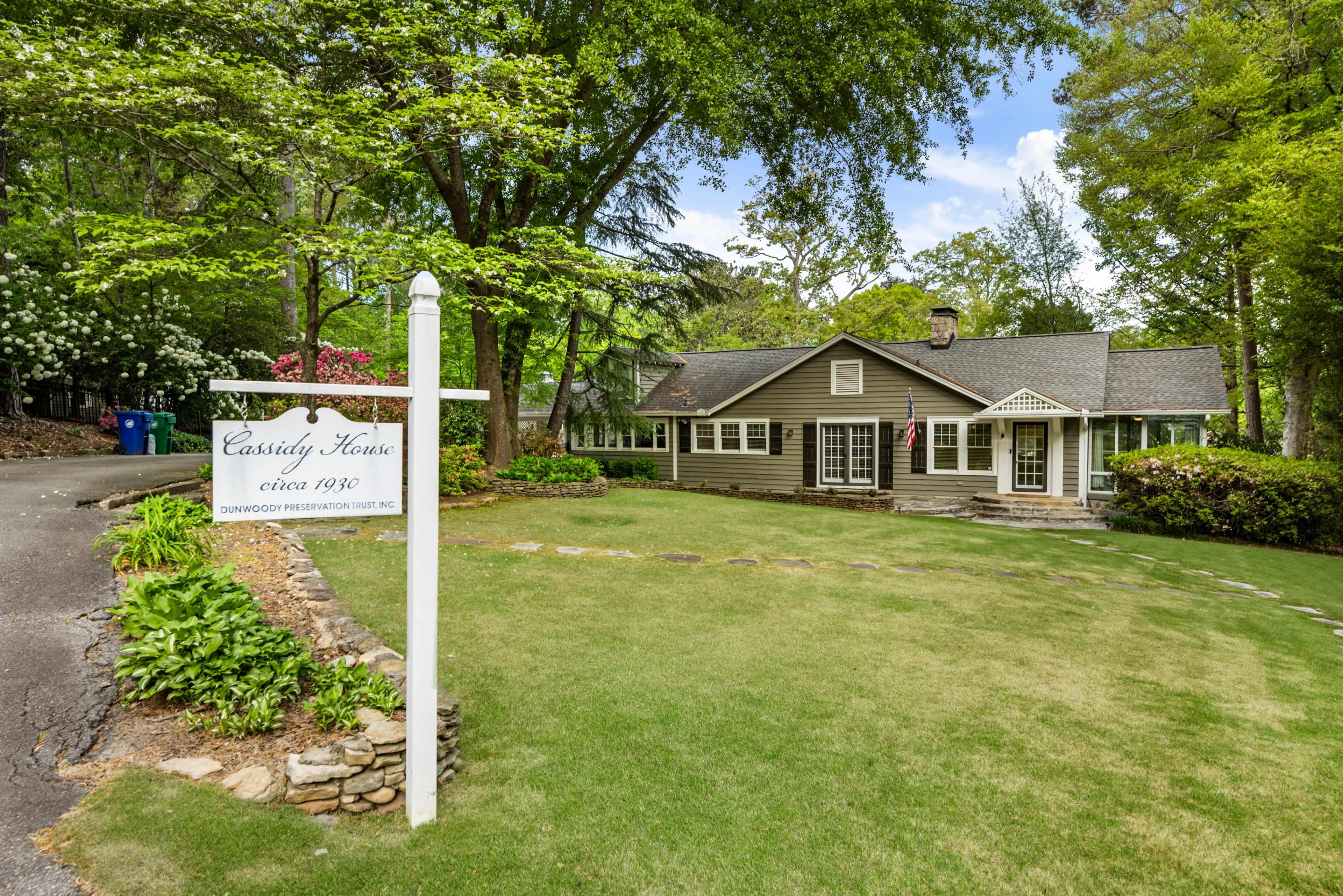 The image shows a gray house with a front porch, surrounded by lush greenery and a stone pathway, along with a sign that reads "Cassidy House circa 1930."