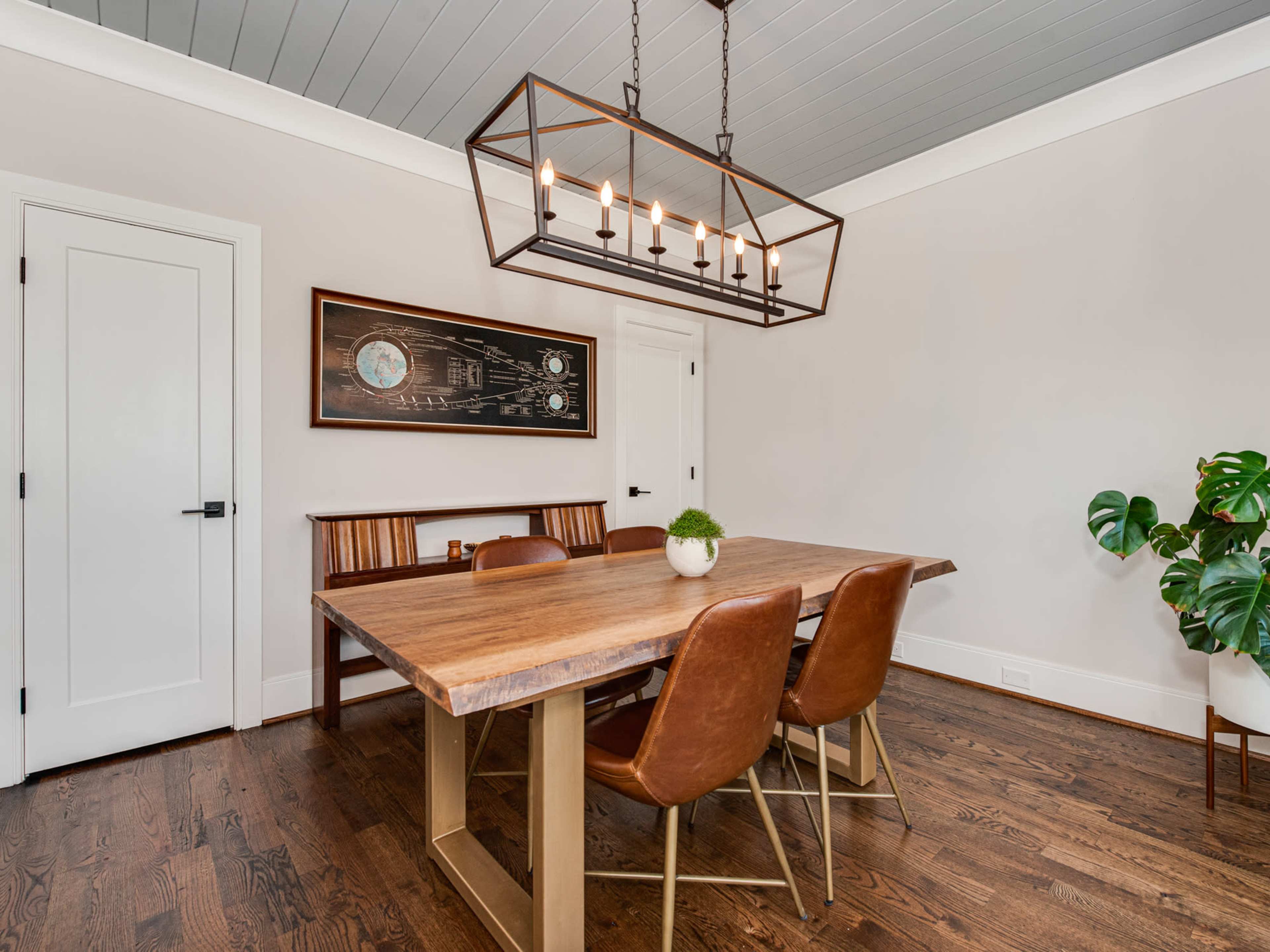 A wooden dining table with leather chairs is positioned under a metal light fixture in a room with a painted ceiling and a framed wall art.