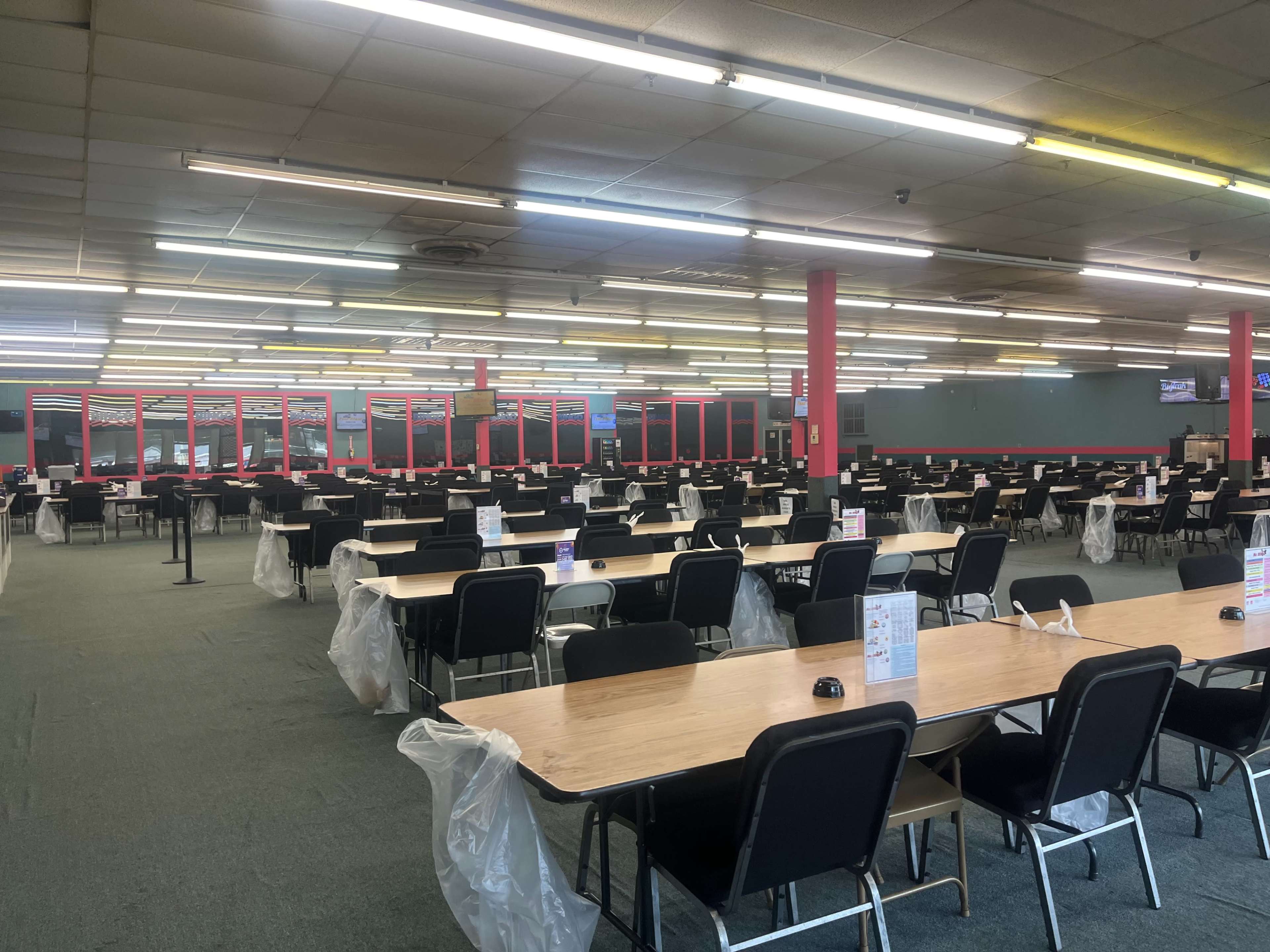 A large, empty dining hall with numerous tables and chairs arranged in rows, illuminated by overhead fluorescent lights.
