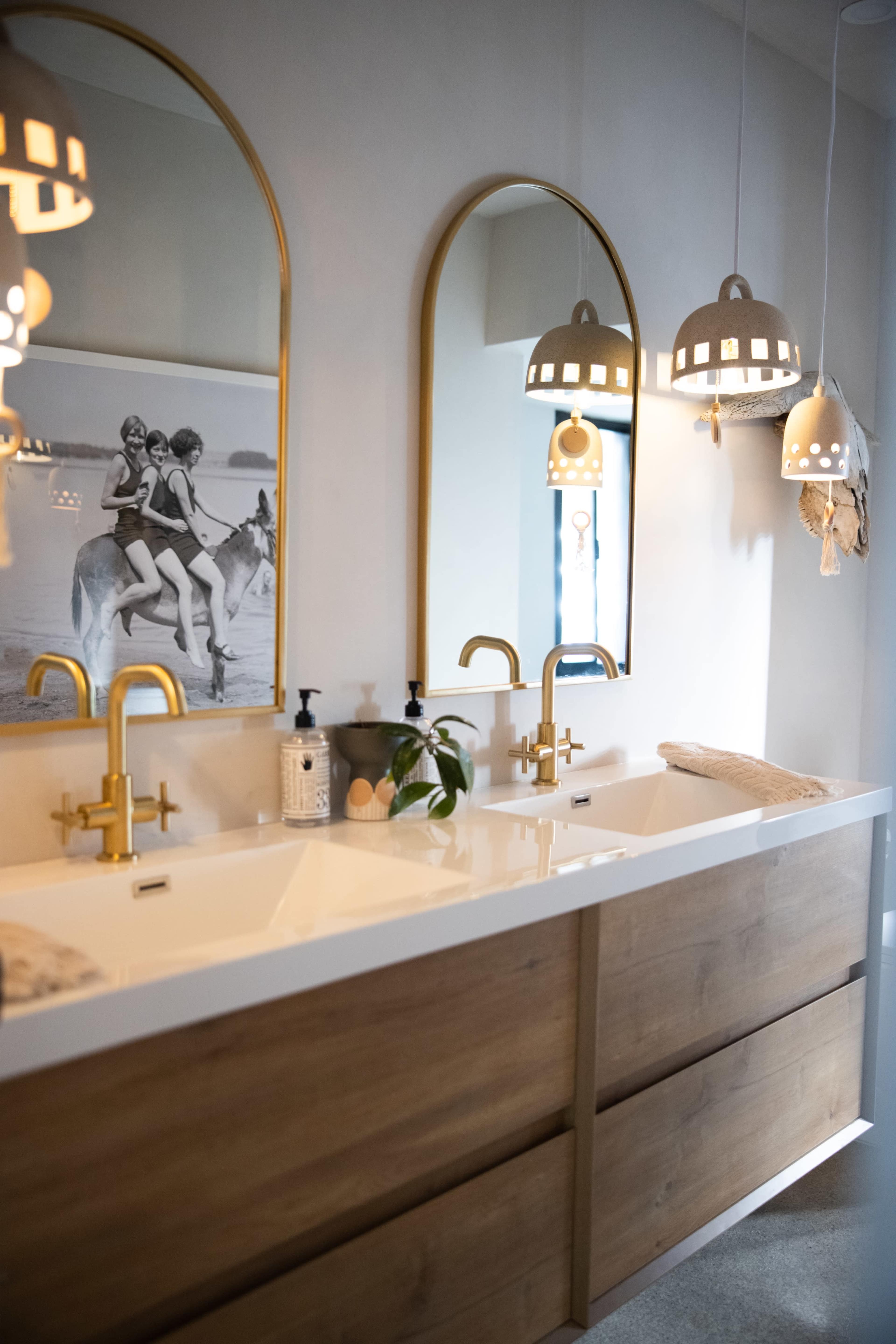 A modern bathroom with two sinks, gold faucets, and large round mirrors, illuminated by unique pendant lights.