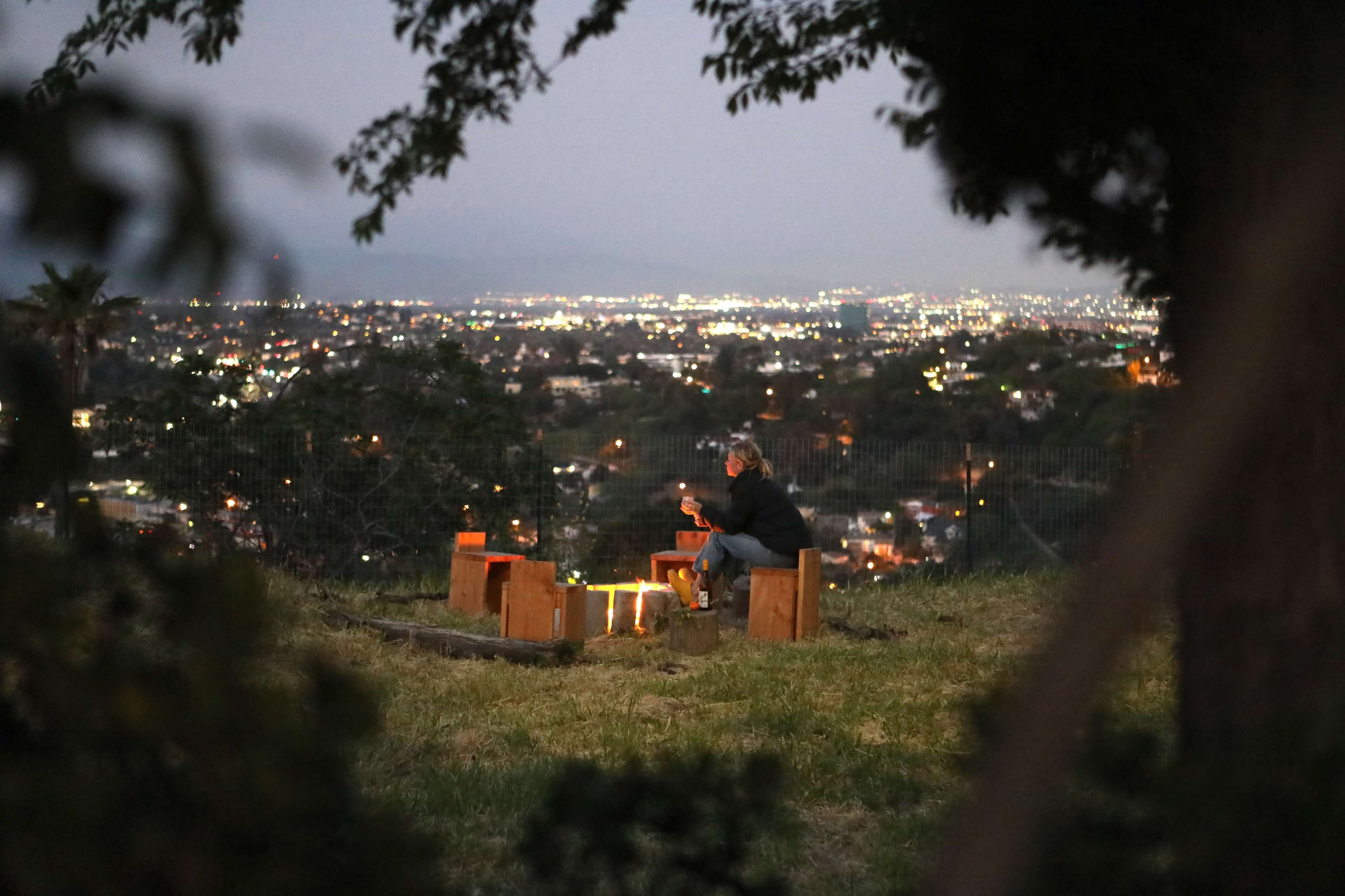 A person sits on benches around a campfire overlooking a city illuminated at night.