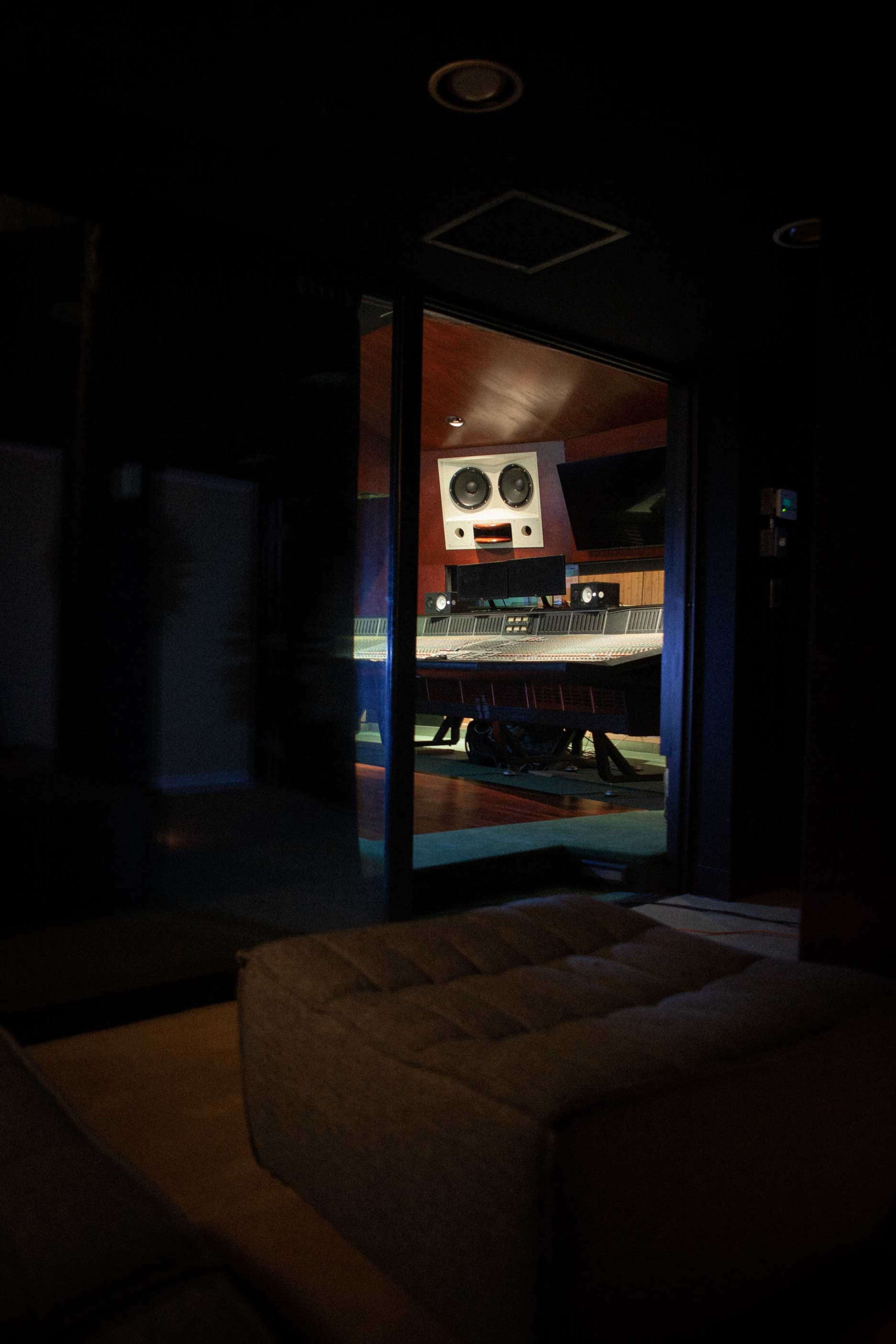 A dimly lit recording studio featuring a large audio mixing console viewed through a glass wall.