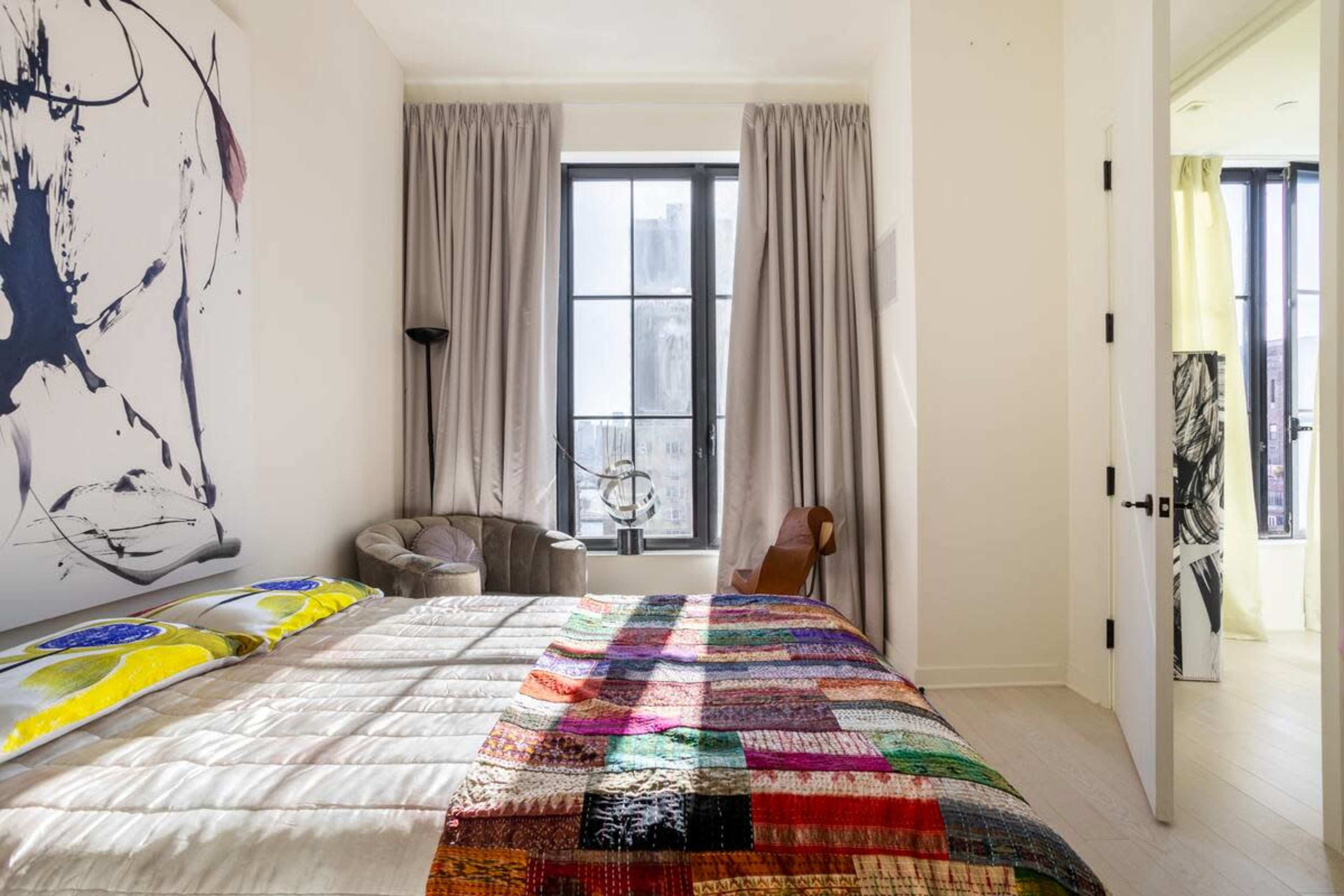 The image shows a modern bedroom featuring a colorful patchwork quilt on the bed, a comfortable chair in one corner, and large windows with light gray curtains.