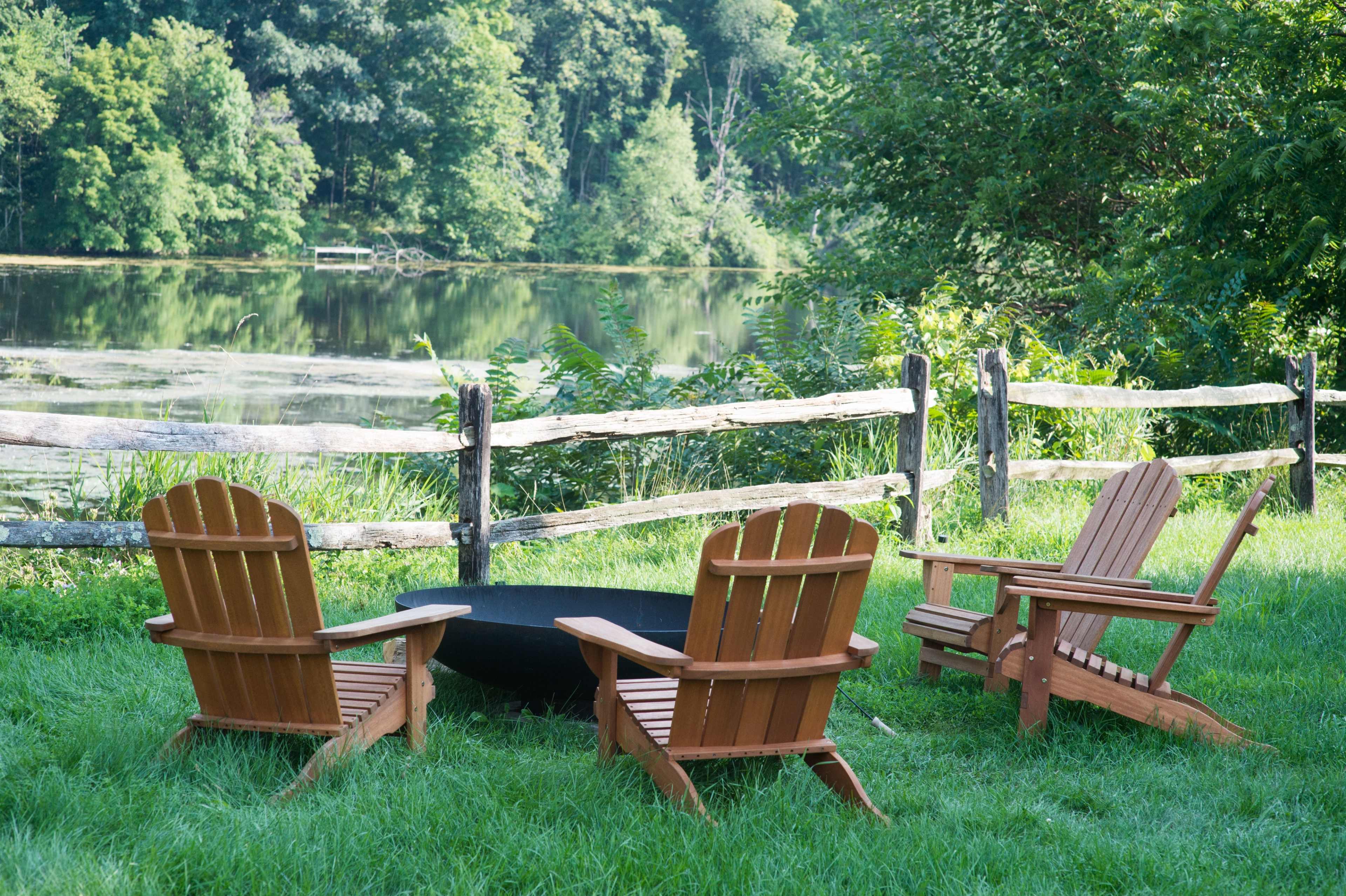 Three wooden Adirondack chairs are arranged around a black fire pit near a peaceful pond surrounded by greenery and a wooden fence.