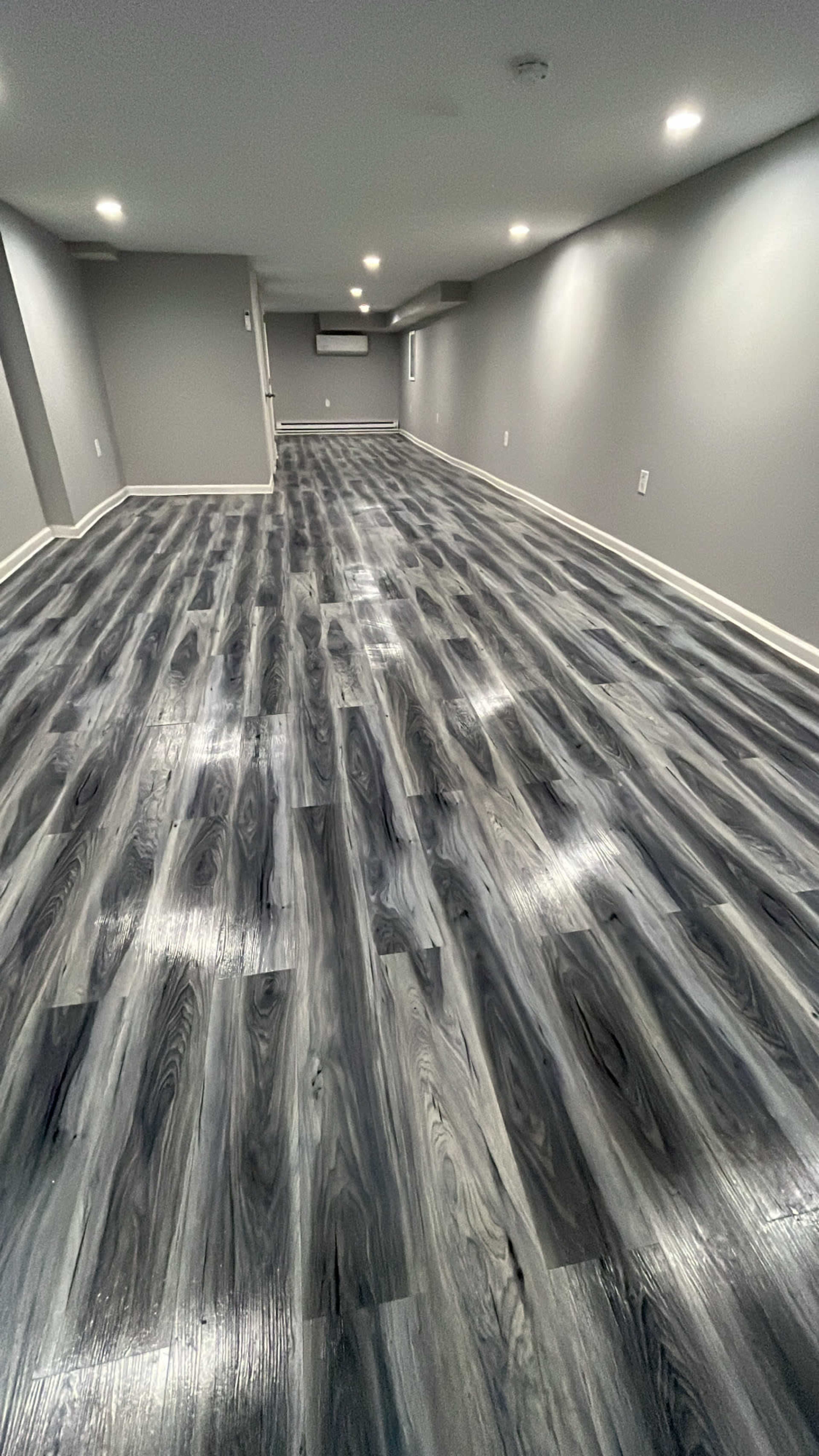 A spacious basement room with gray walls and dark wood-like flooring illuminated by overhead lights.