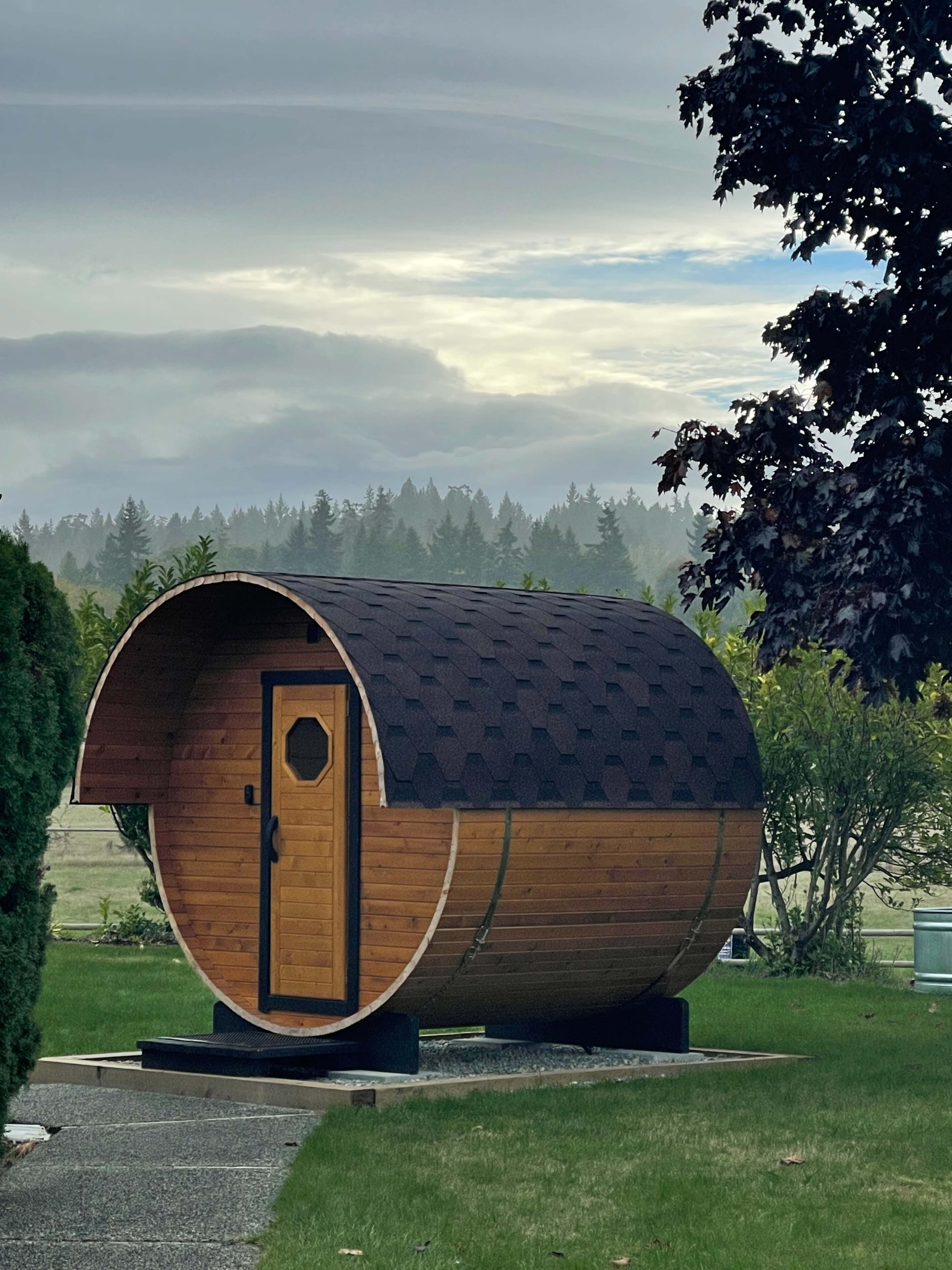 A wooden barrel-shaped sauna stands on a grassy area with trees and a cloudy sky in the background.