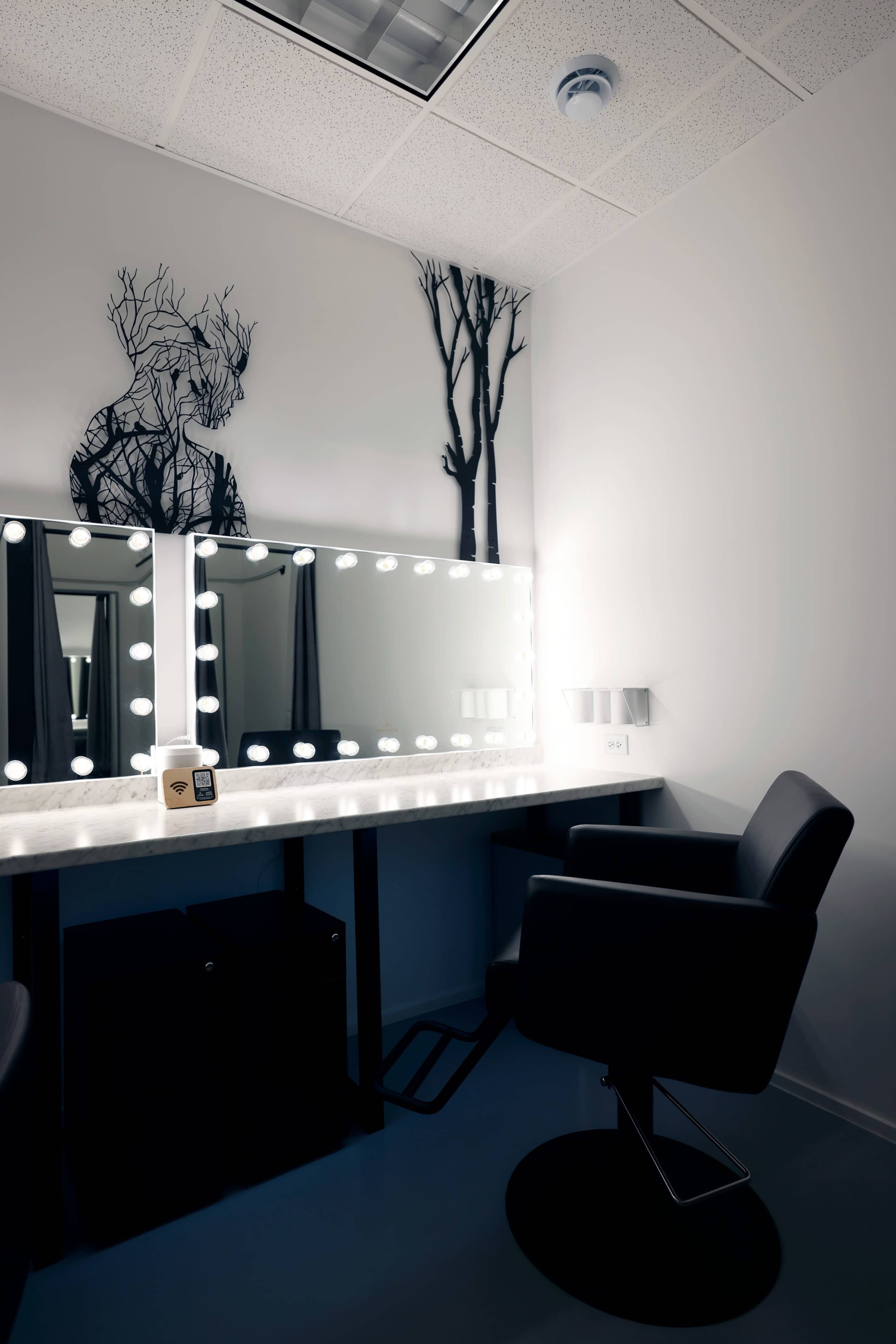 A minimalist dressing room with a illuminated mirror, a chair, and a decorative tree design on the wall.