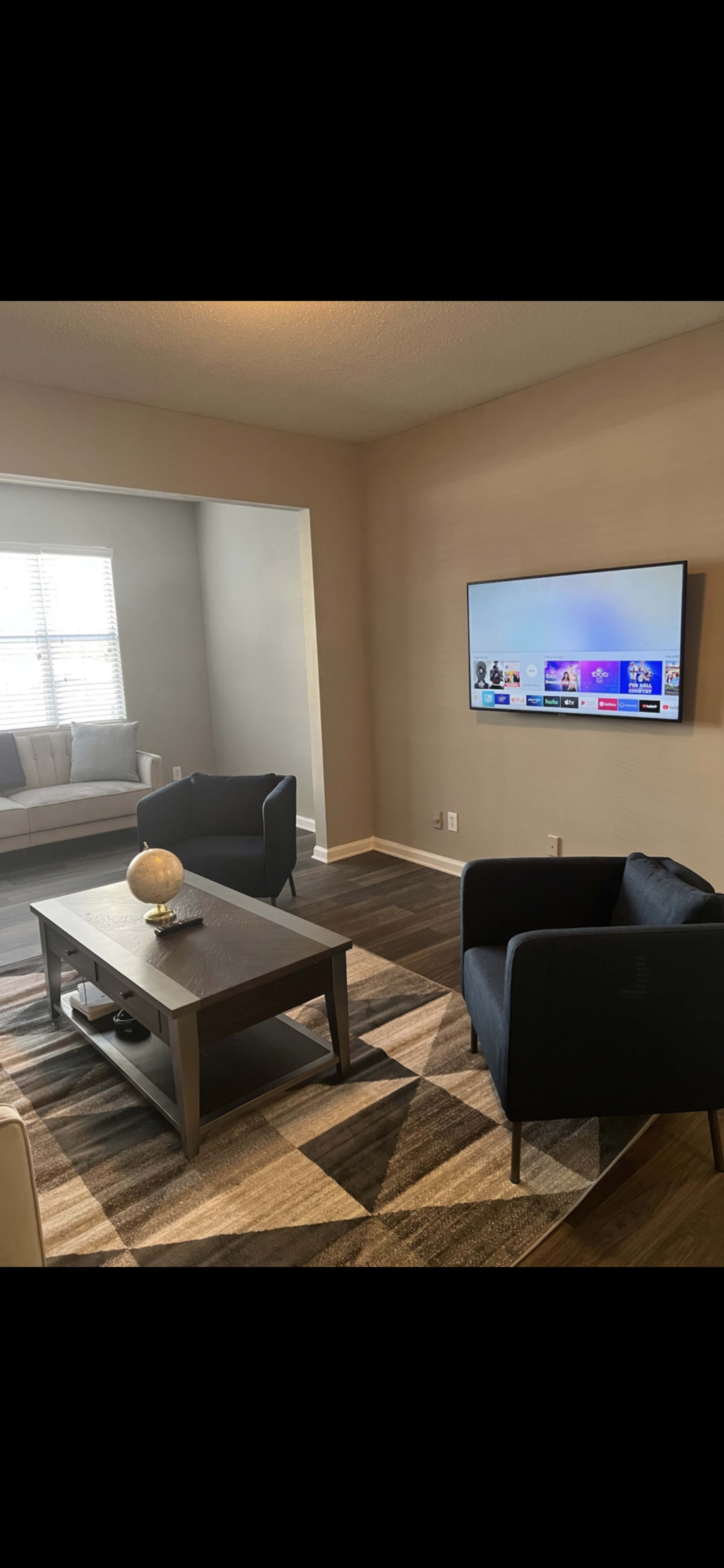 A living room with a television mounted on the wall, a coffee table in the center, and two chairs facing each other on a patterned rug.