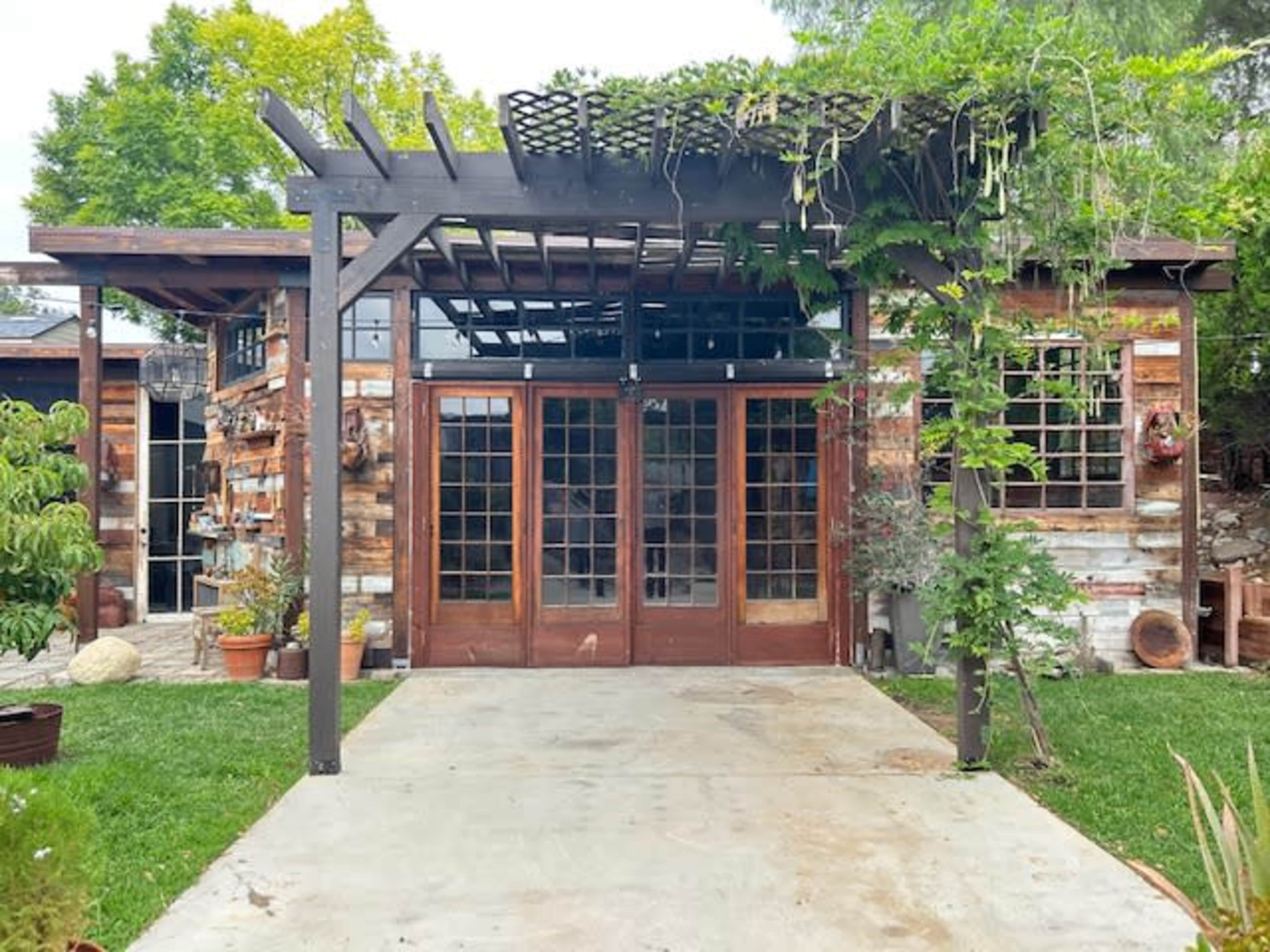 A rustic wooden structure with large glass doors, surrounded by greenery and a pergola with climbing plants.