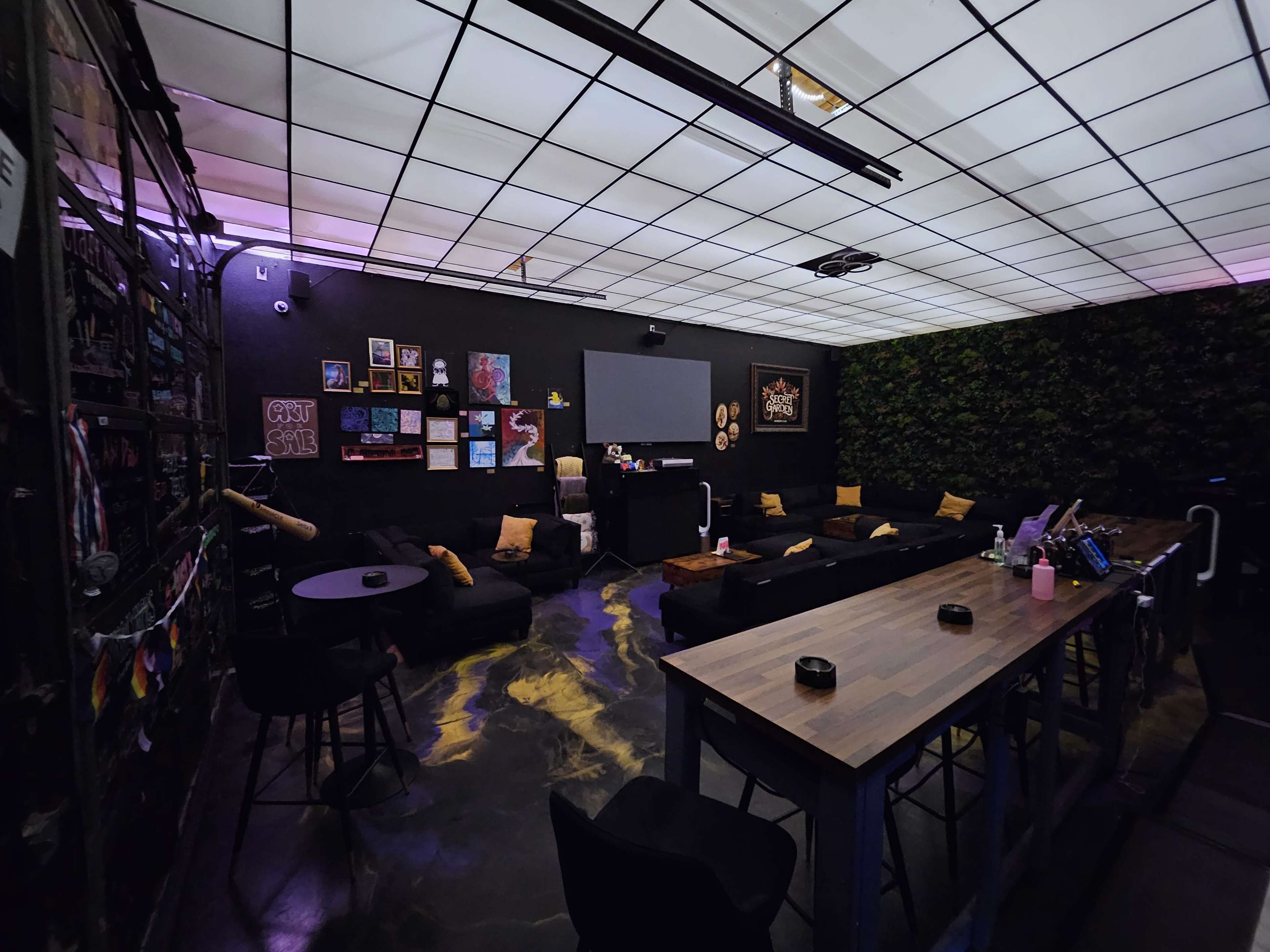 The image shows a modern lounge area with black walls, a large screen on one side, and various seating options arranged around a wooden table.