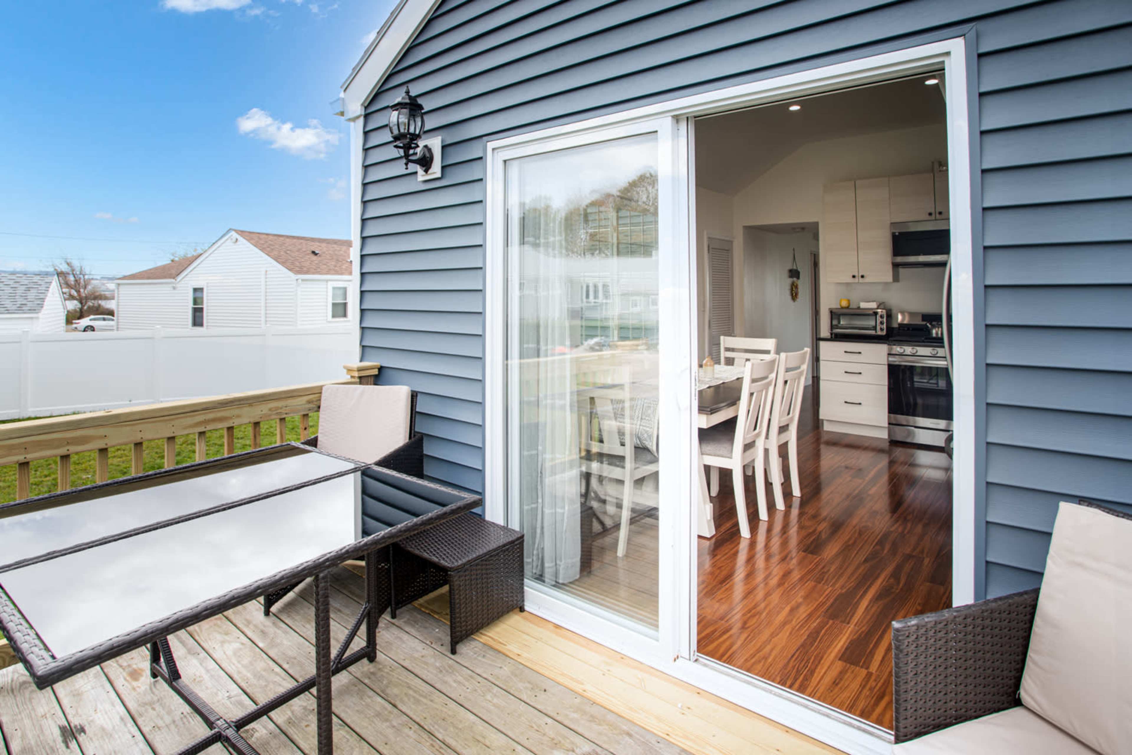 A sliding glass door opens from a wooden deck into a bright kitchen with white furniture.