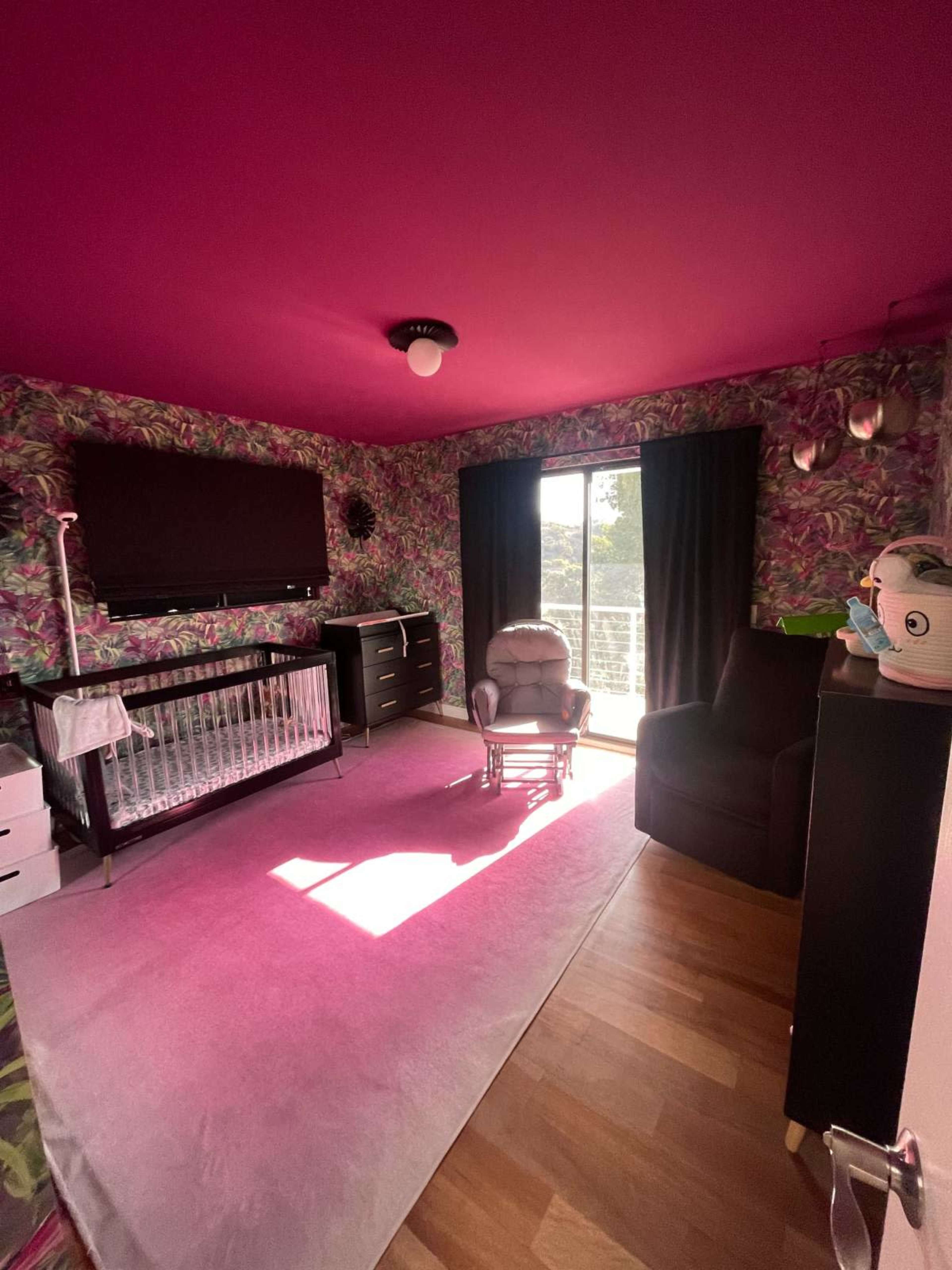 The image shows a brightly decorated nursery with a pink ceiling, floral wallpaper, a crib, a rocking chair, and a large window letting in natural light.