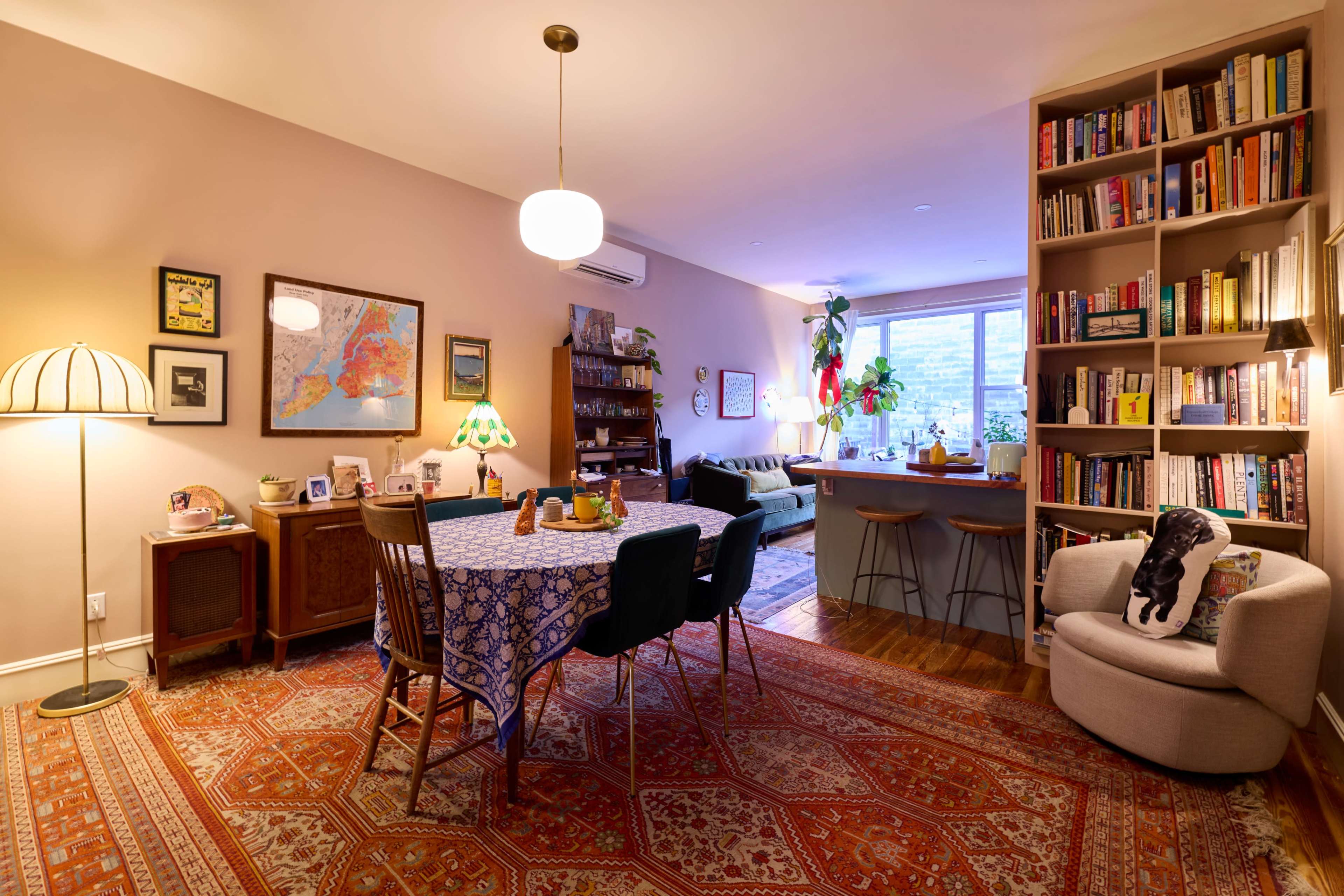 The image shows a cozy living space featuring a dining area with a patterned tablecloth, surrounded by chairs, a bookshelf filled with books, a sofa in the background, and warm lighting.