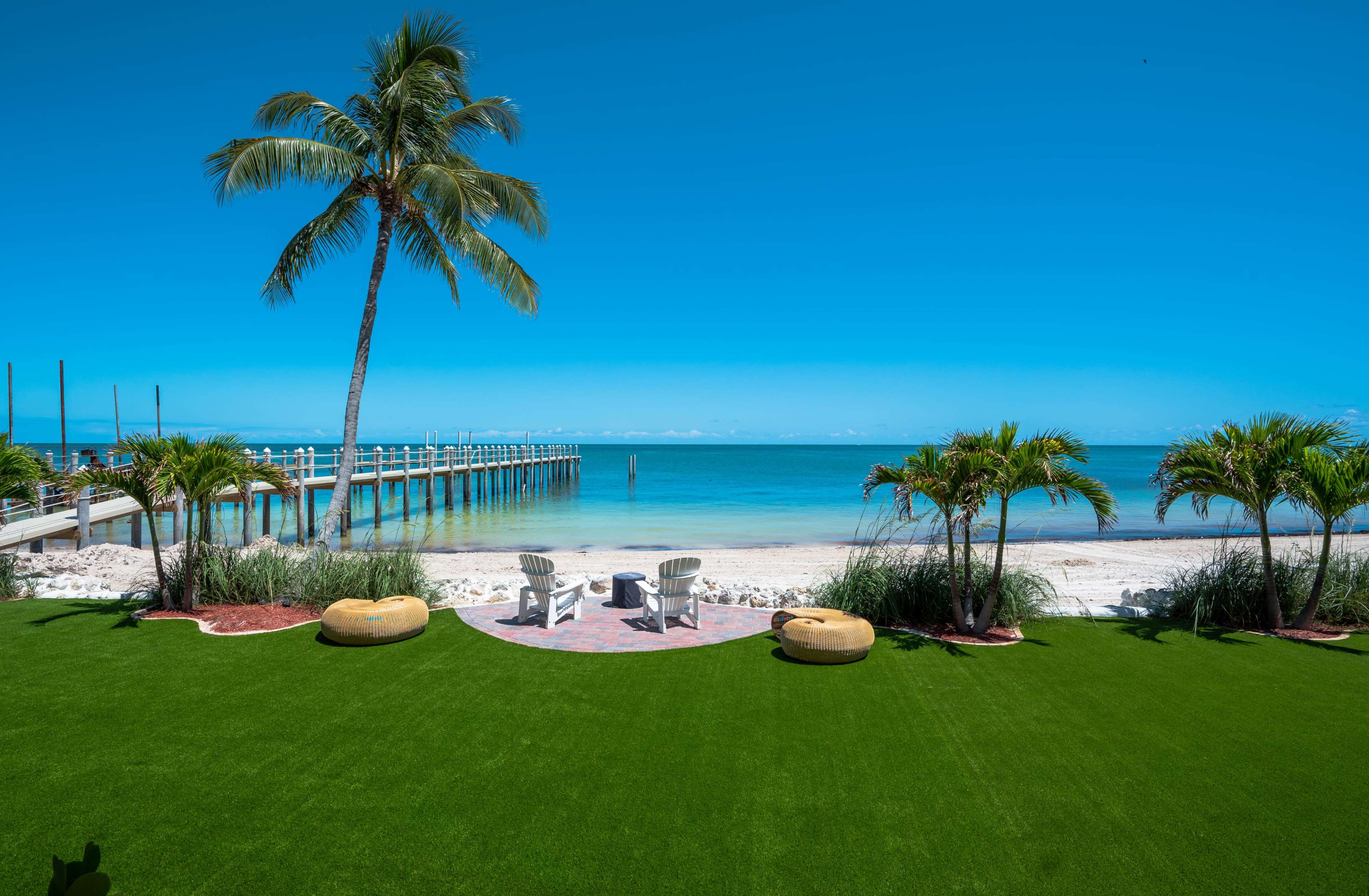 A sandy beach with a wooden pier and two chairs facing the ocean, framed by palm trees and green landscaping.