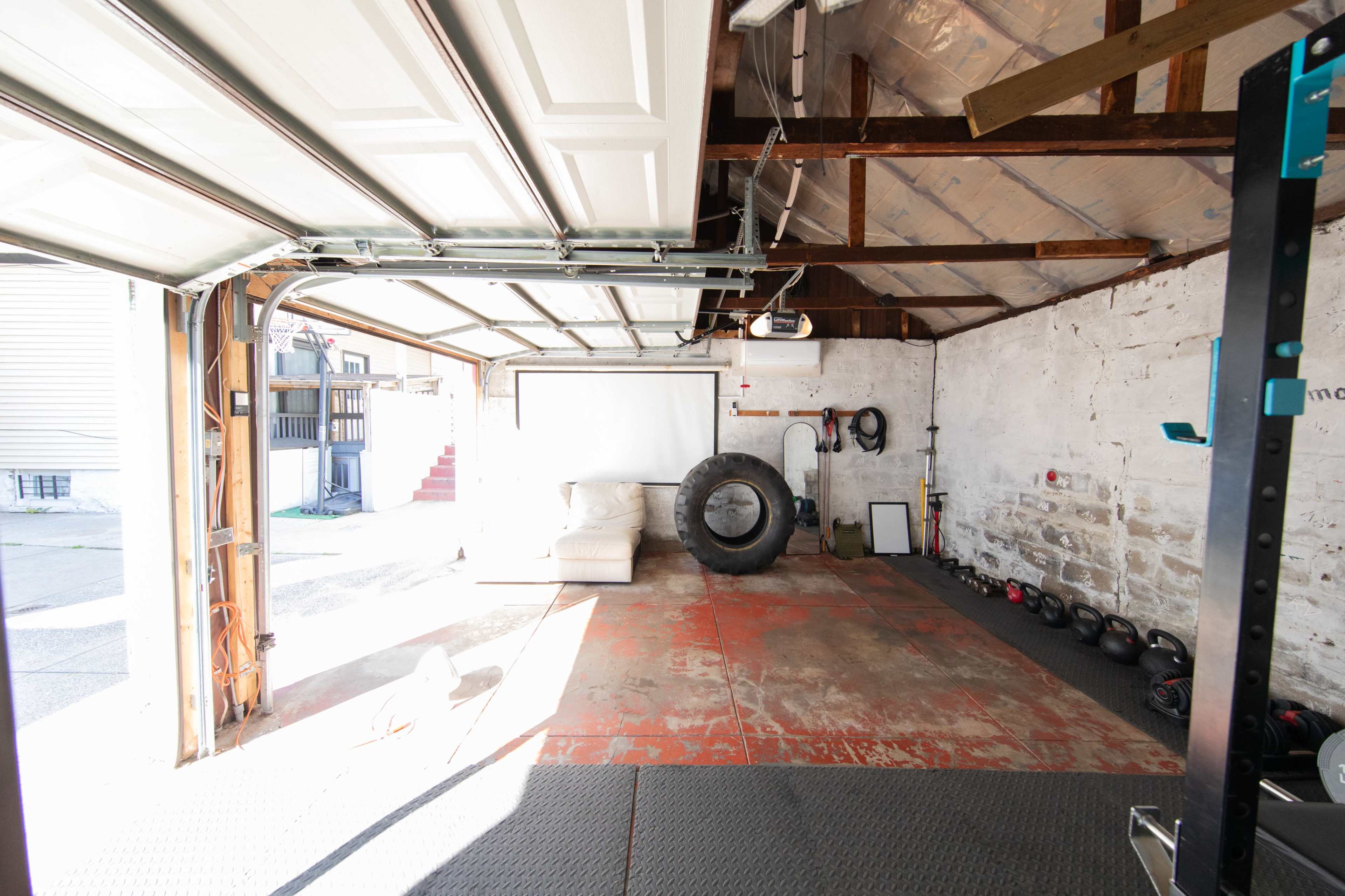 The image shows a garage gym with a sofa, a large tire, exercise equipment, and an open garage door letting in natural light.