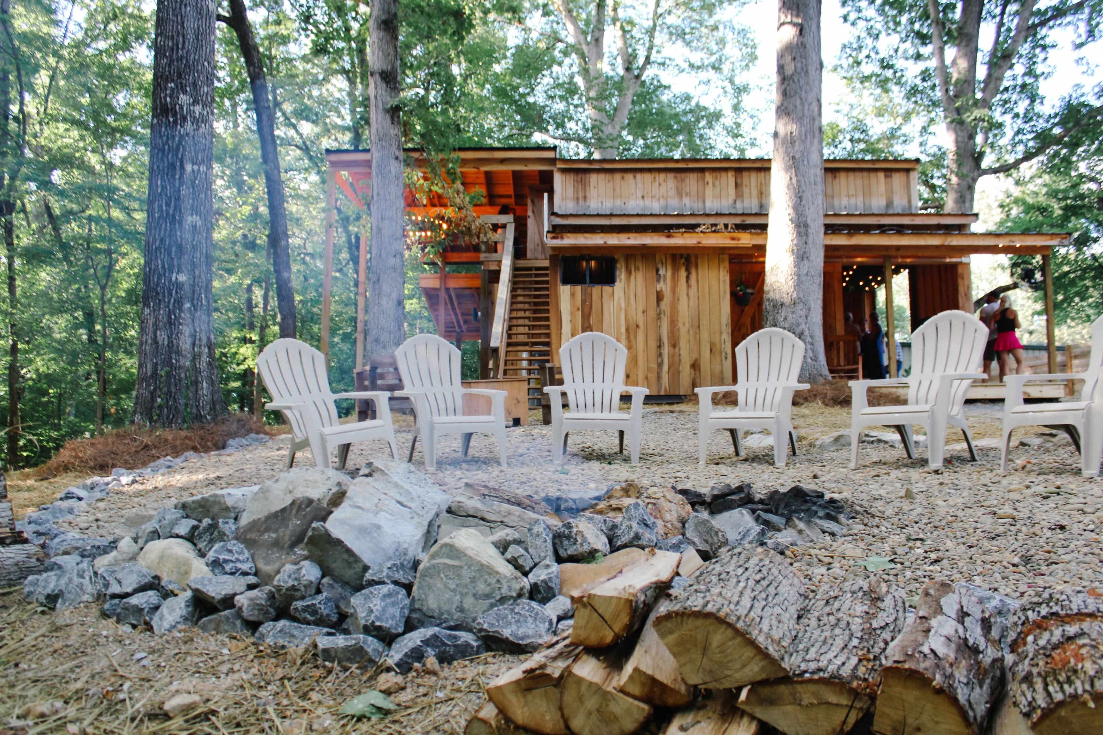 A rustic wooden cabin surrounded by trees features a fire pit with several white chairs arranged in a circular formation.
