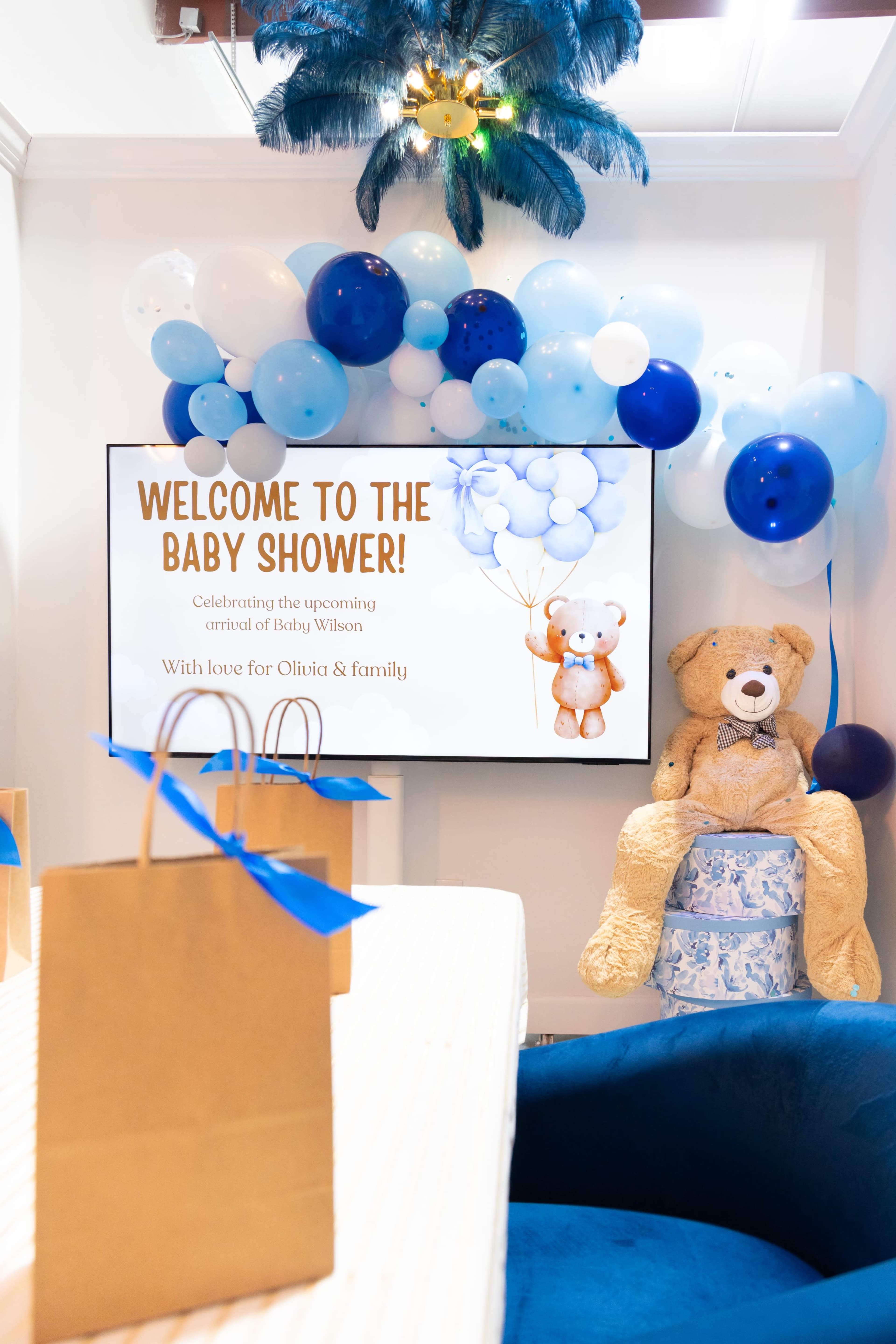 The image shows a decorated room for a baby shower, featuring a screen with a welcome message, blue balloons, a teddy bear, and gift bags on a table.