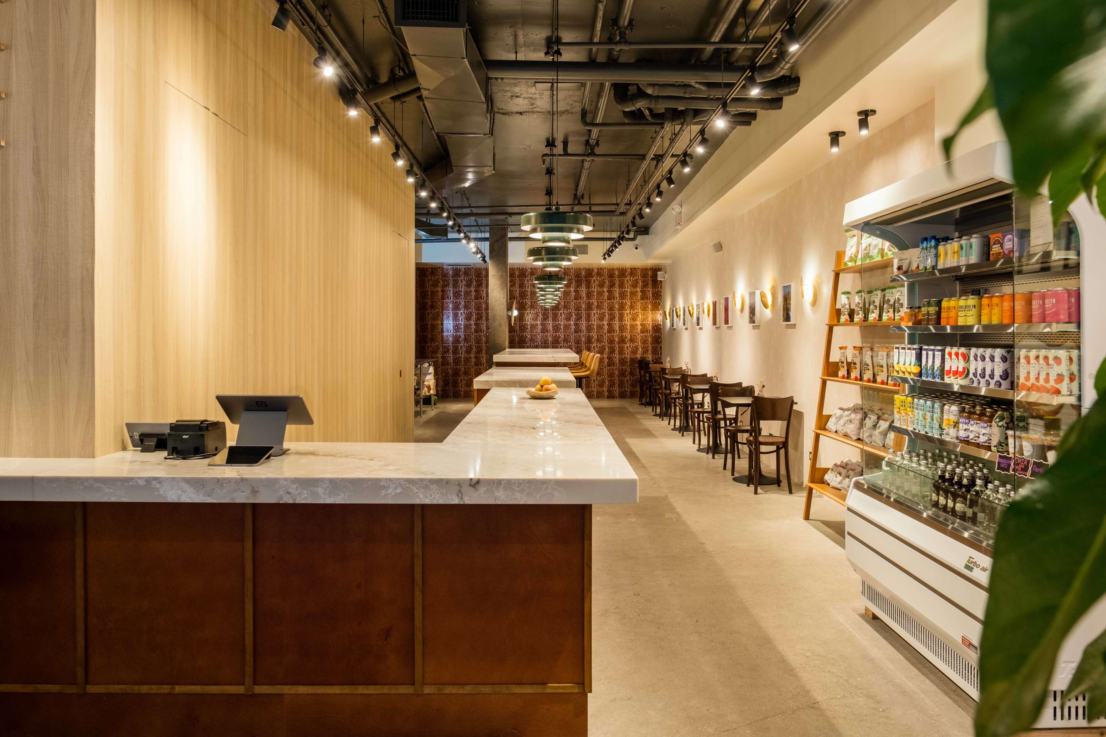 The image shows a modern cafe interior featuring a long, empty counter with seating on one side and shelves displaying beverages and snacks on the other.