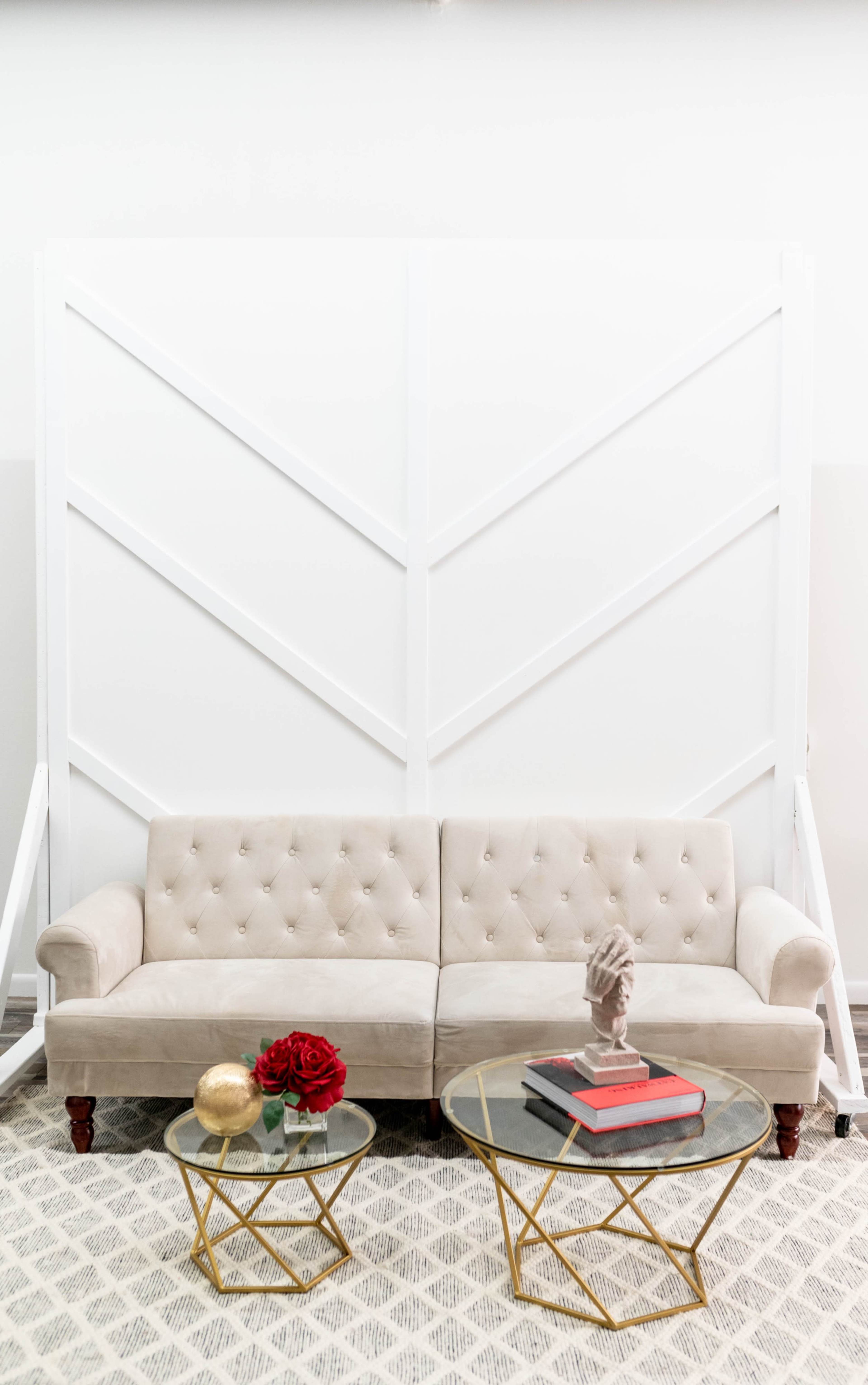 A cream-colored, tufted sofa positioned in front of a geometric white backdrop, with two glass coffee tables adorned with books and decorative items.