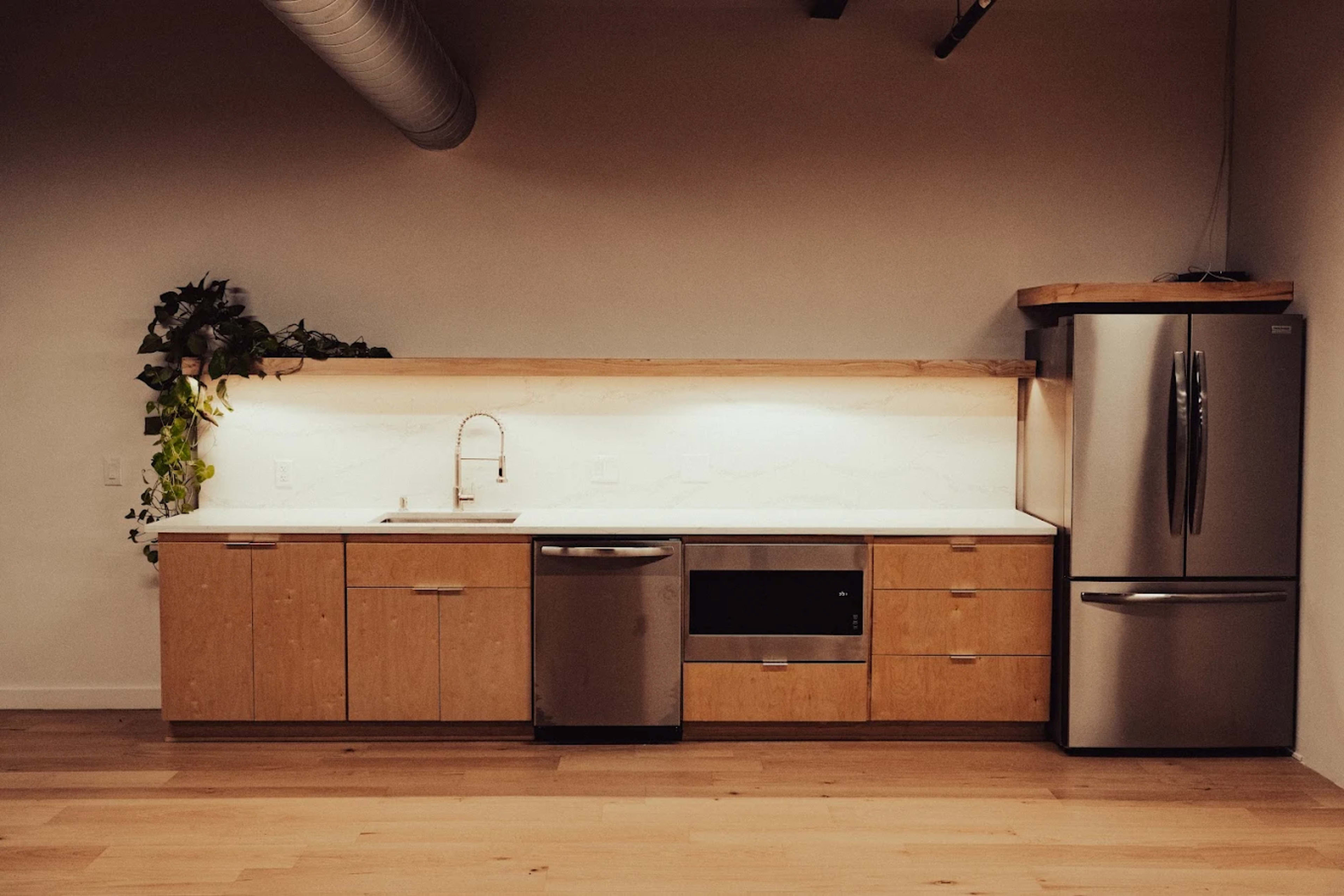 The image shows a modern kitchen with wooden cabinets, stainless steel appliances, and a white countertop, illuminated by under-cabinet lighting.