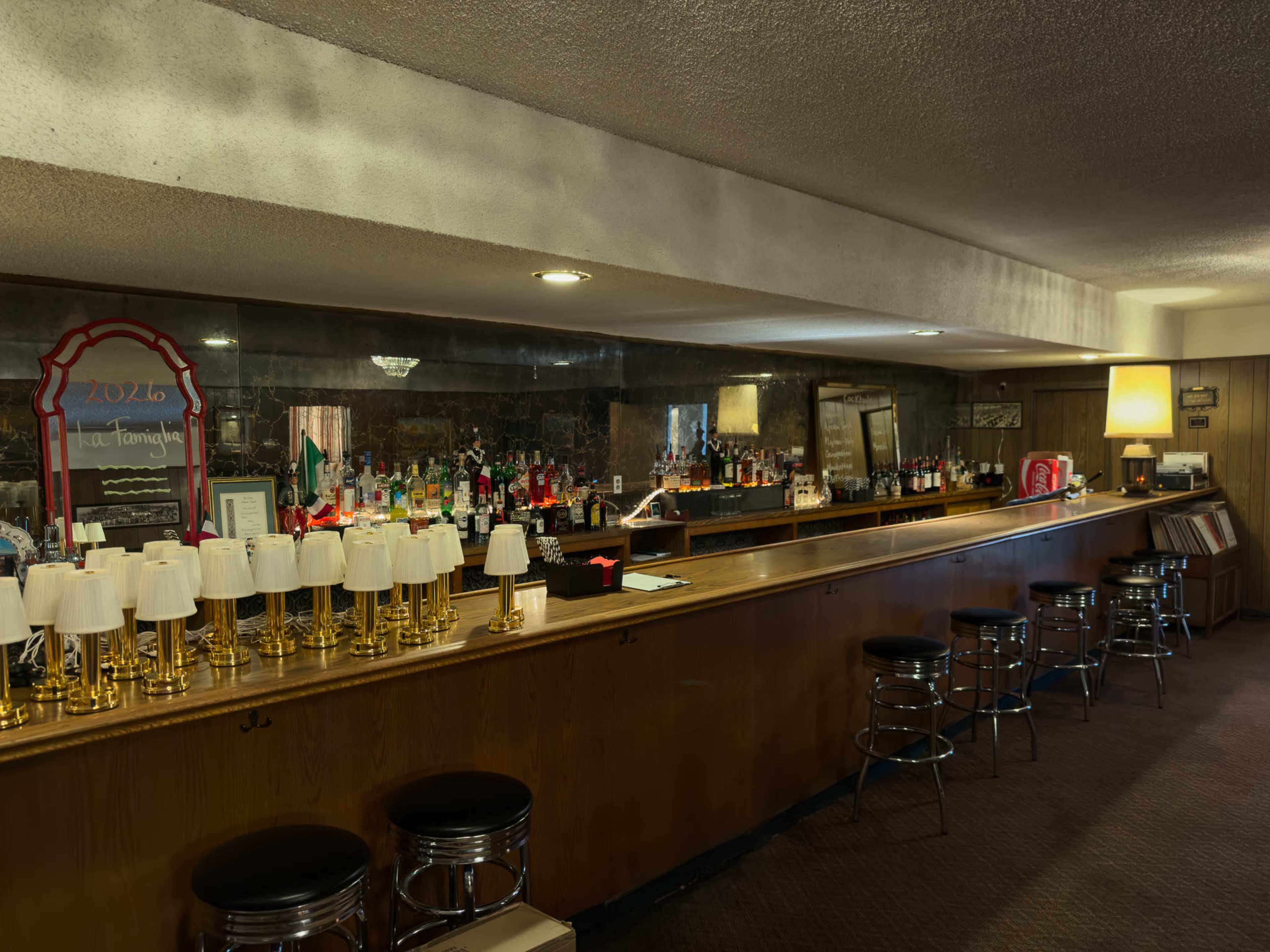 A dimly lit bar features a long wooden counter with several lighted glass fixtures and a mirrored backdrop showcasing various alcohol bottles.