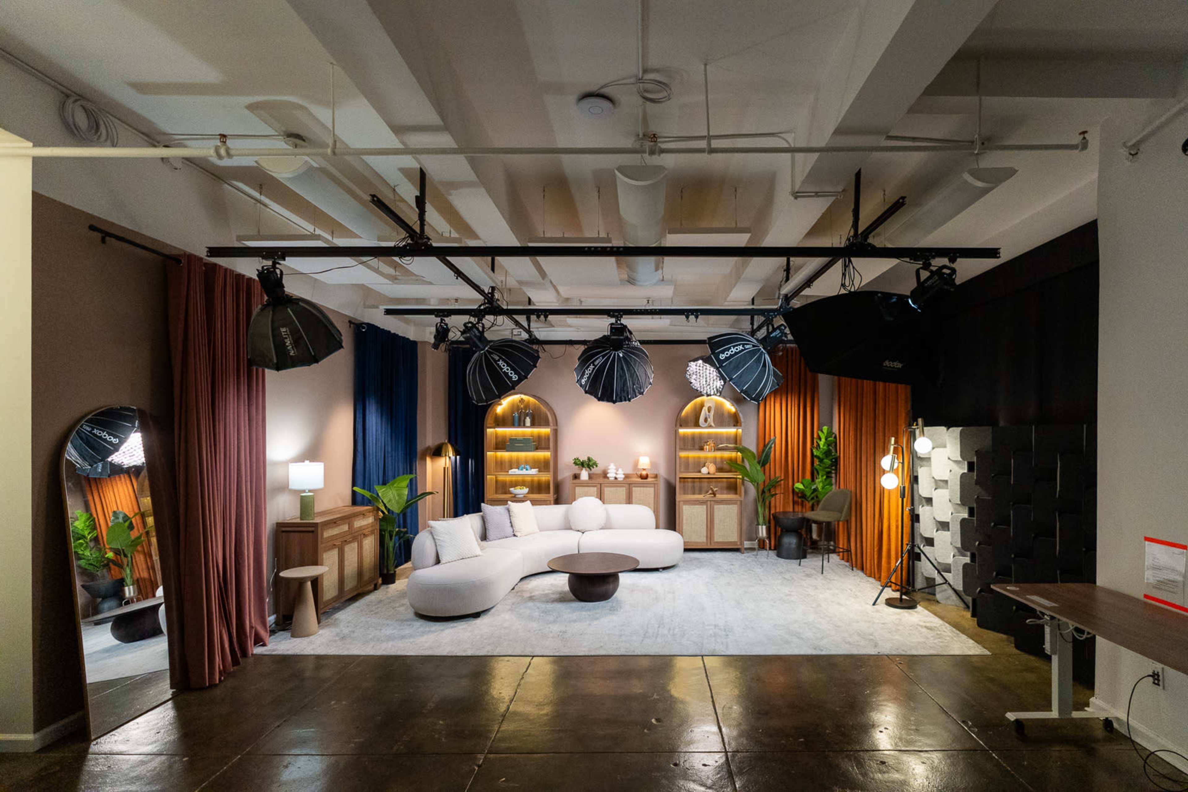 The image shows a well-designed studio space featuring a modern seating area, decorative lighting, and various plants against a backdrop of colored curtains.
