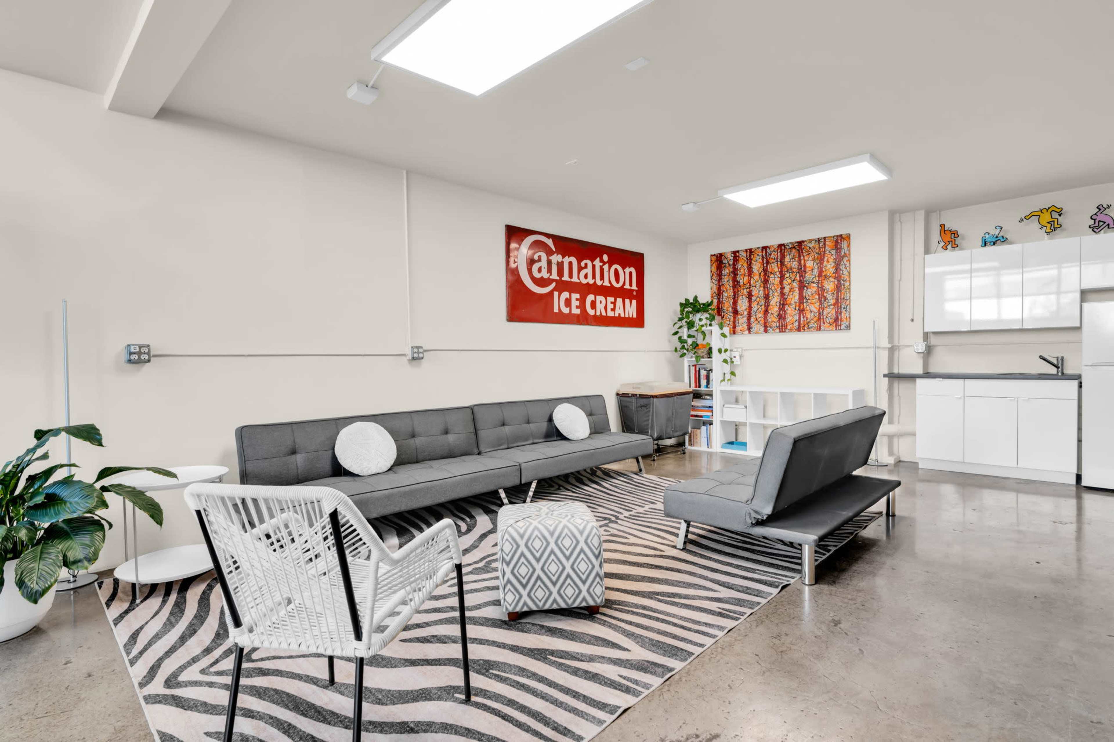 The image shows a modern living space featuring a gray sofa, a white chair, and a kitchenette, with a Carnation Ice Cream sign on the wall.