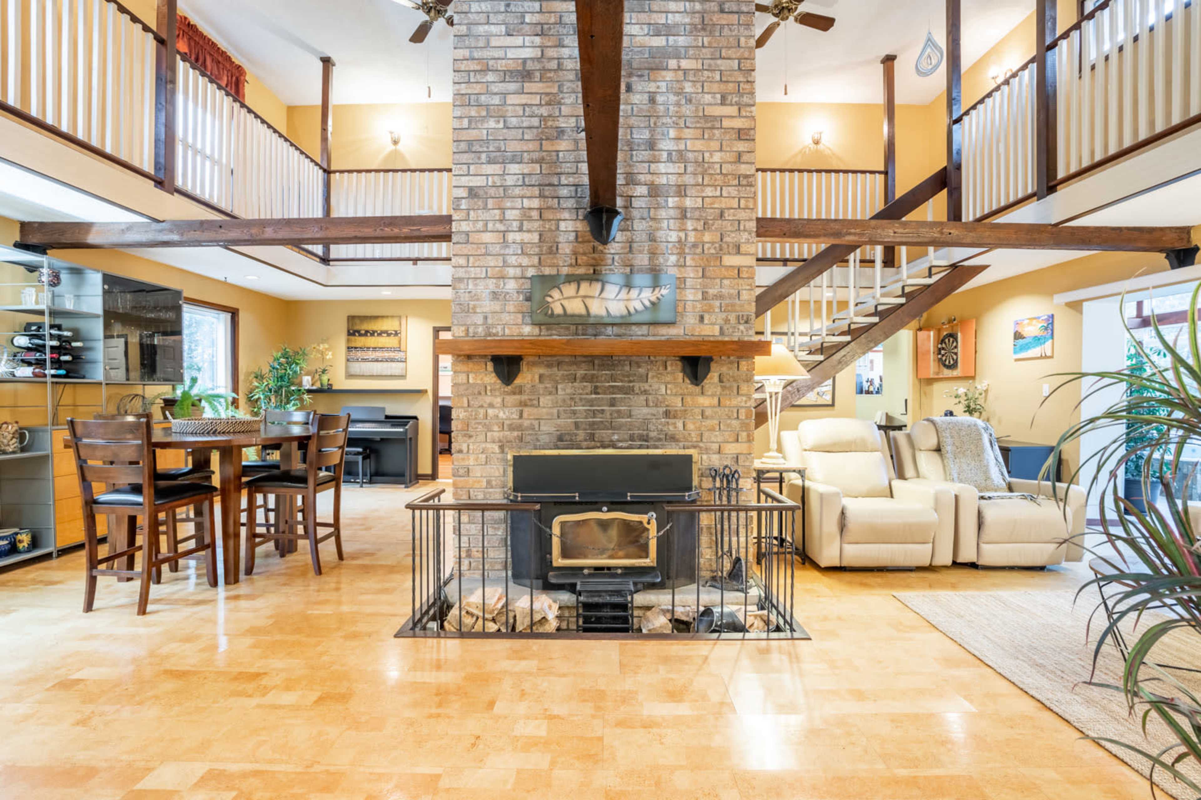 The image shows a spacious living area with a central brick fireplace surrounded by seating, a dining table, and a staircase leading to an upper level.