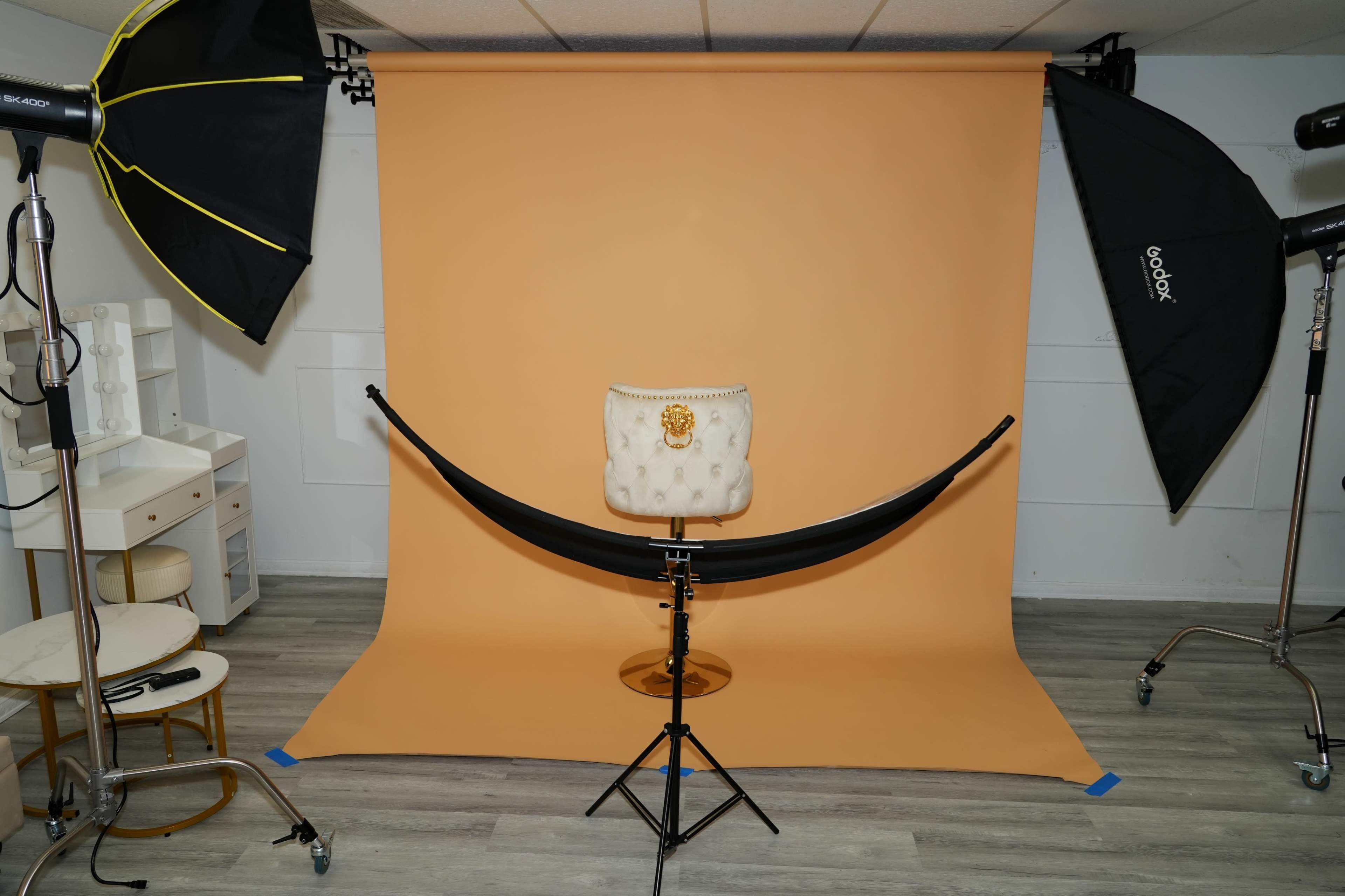 A photography studio setup with a light brown backdrop, a chair in the center, and studio lights positioned on either side.