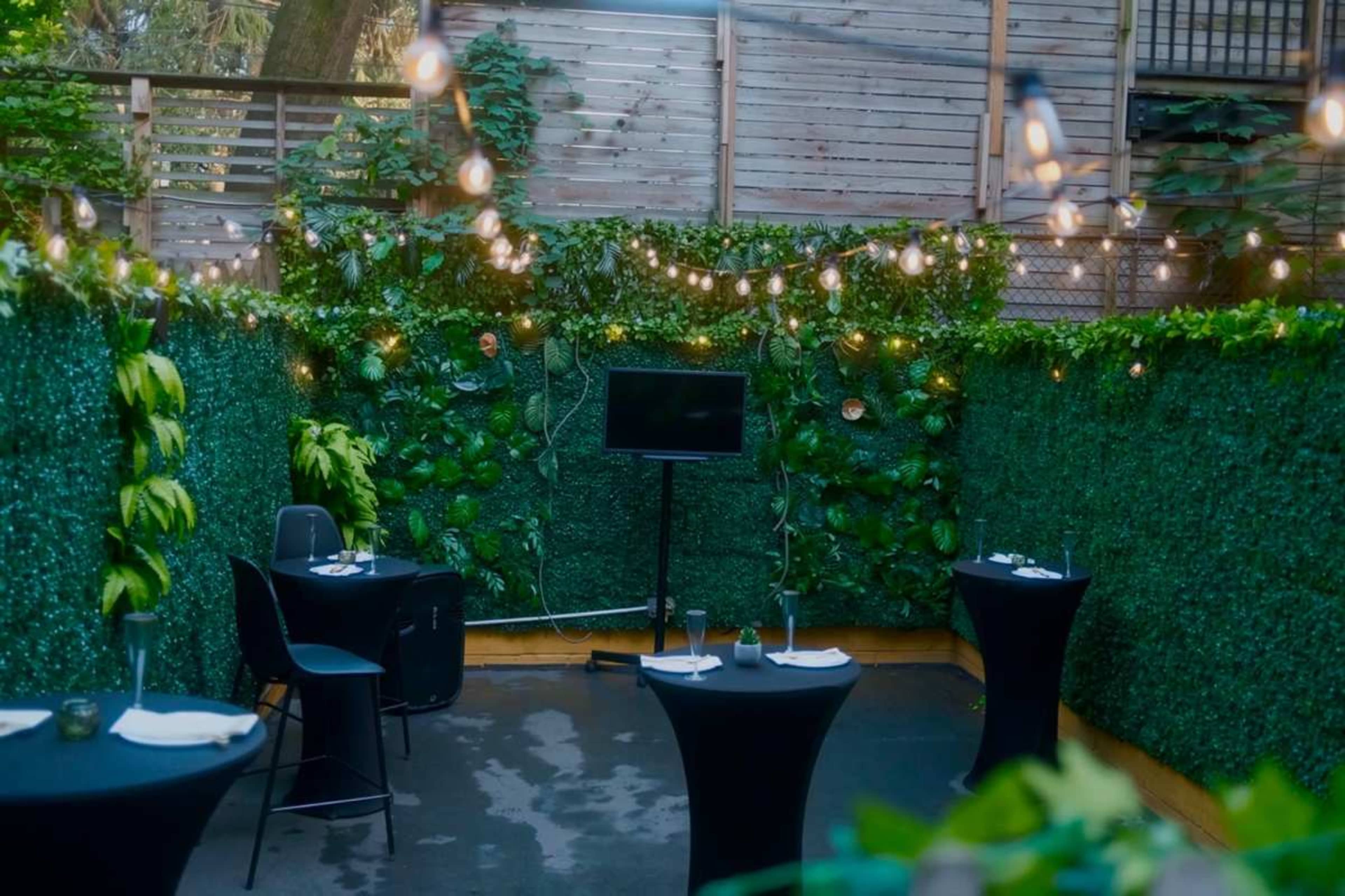 The image shows a small outdoor dining area enclosed by green foliage and string lights, featuring two round black tables set for guests.