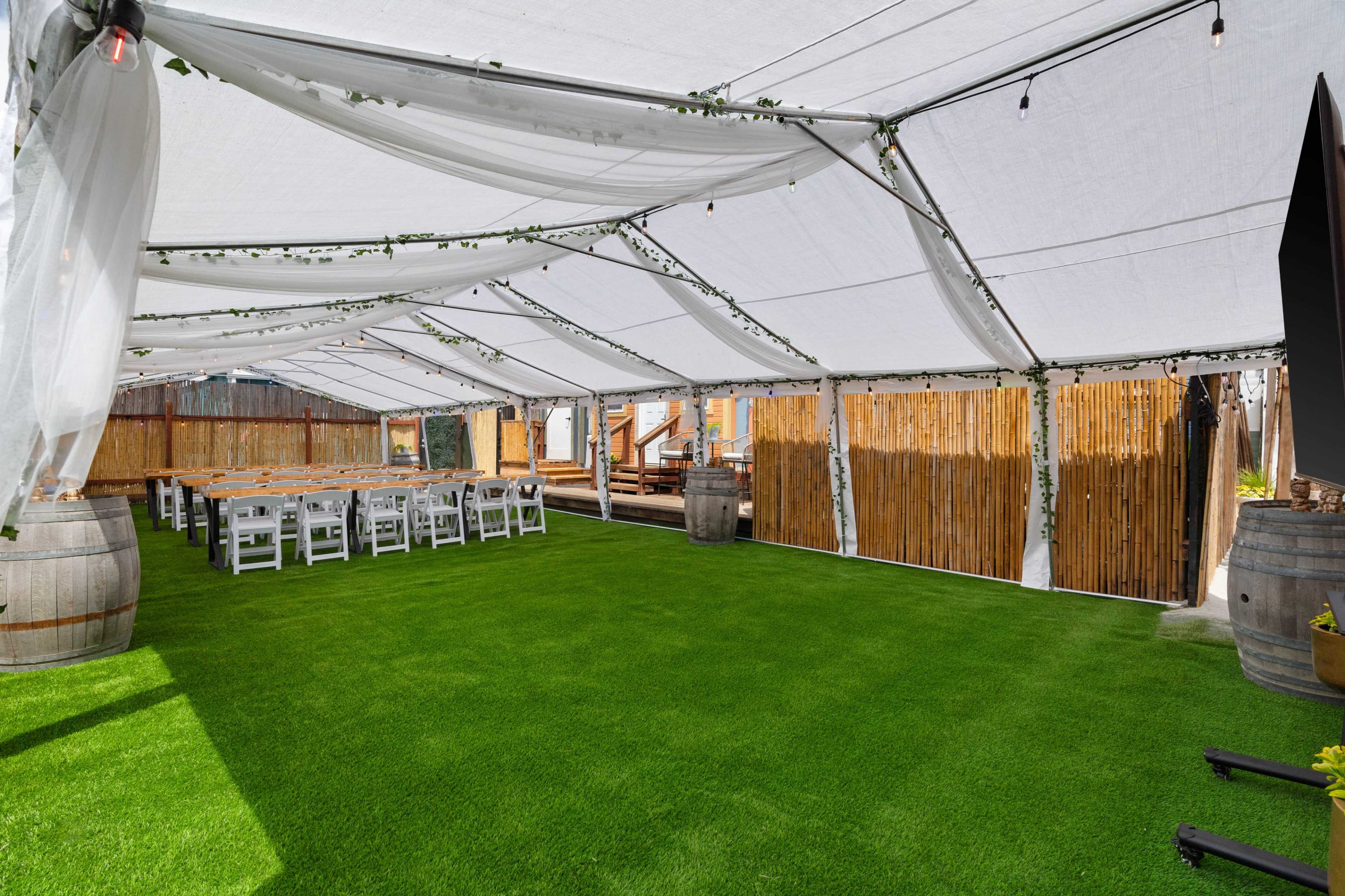 An outdoor event tent features a green carpet, wooden tables set for seating, and barrels as decorative elements.
