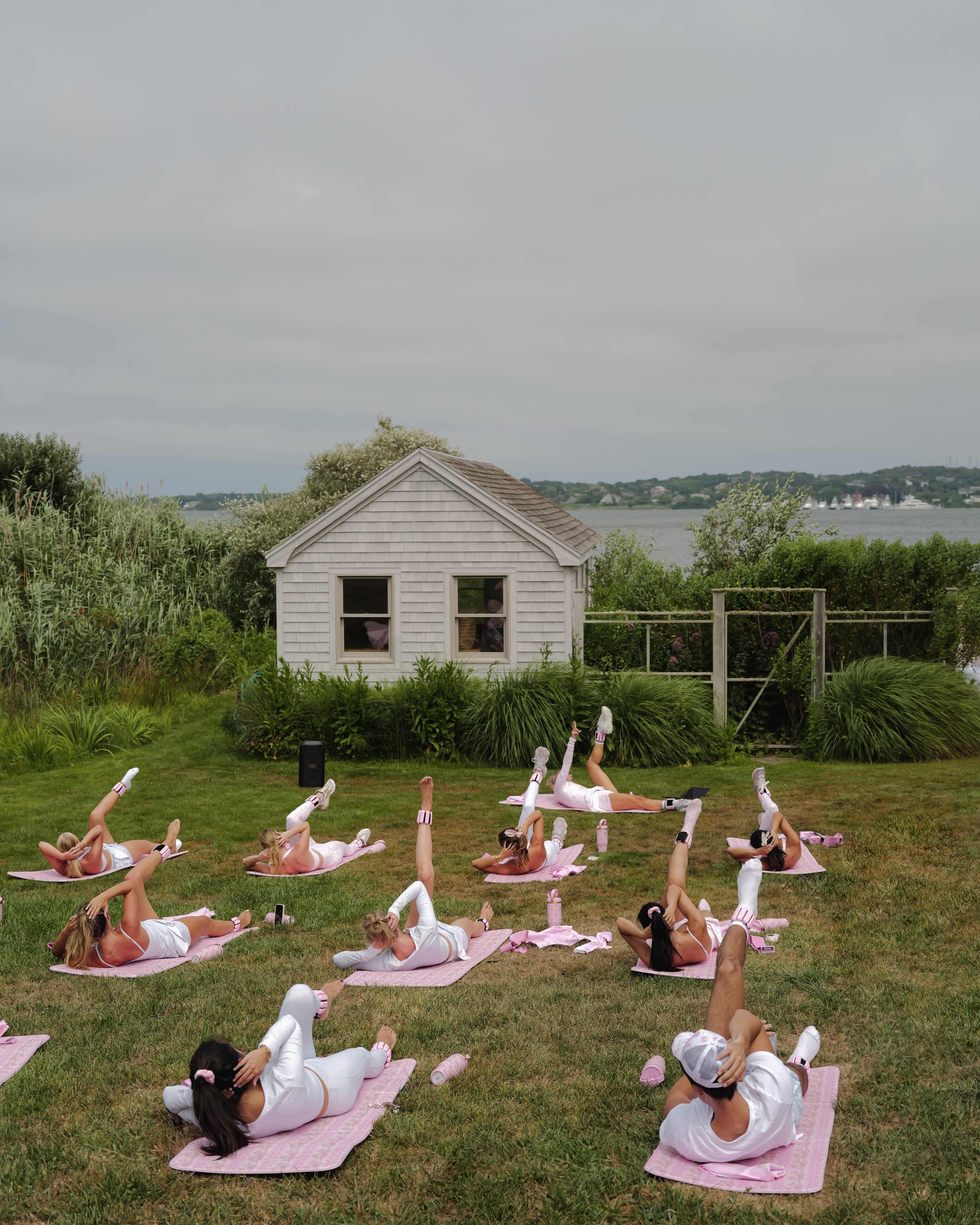 A group of individuals performs leg lifts on yoga mats in a grassy area near a small house and water in the background.